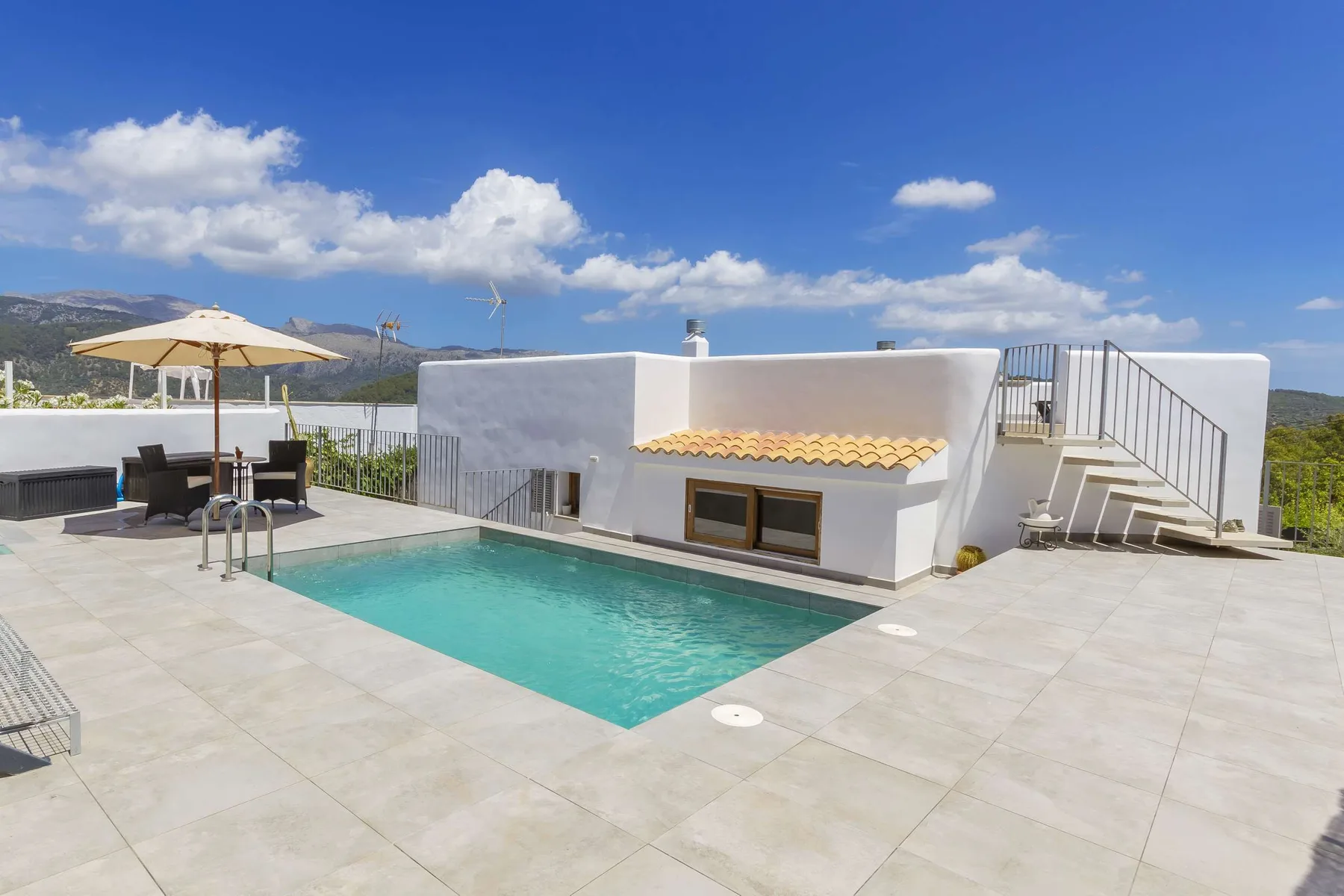A rooftop pool sits beside a white building, with outdoor seating and hills beyond.