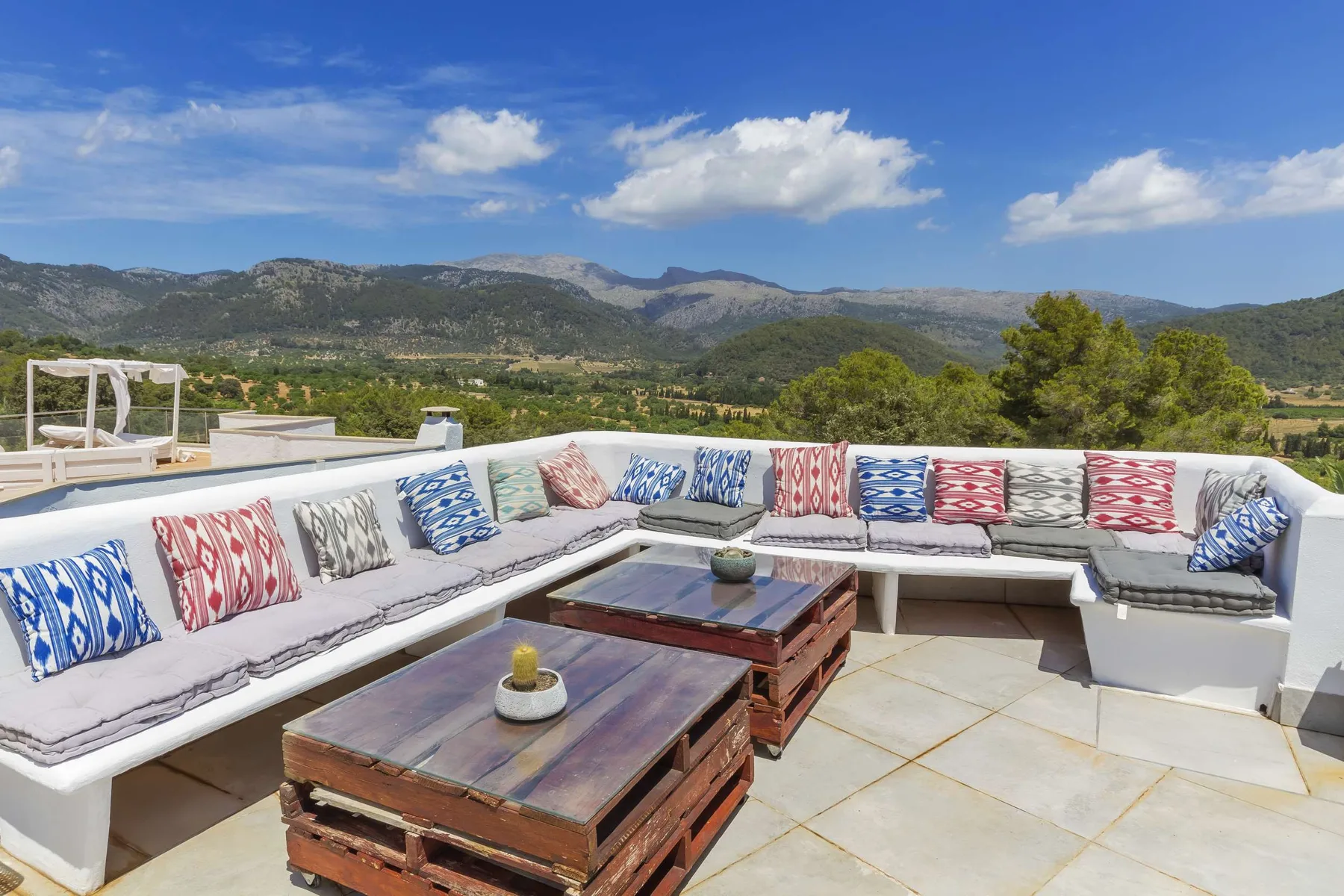 An outdoor rooftop seating area has cushioned benches and wooden tables overlooking hills.