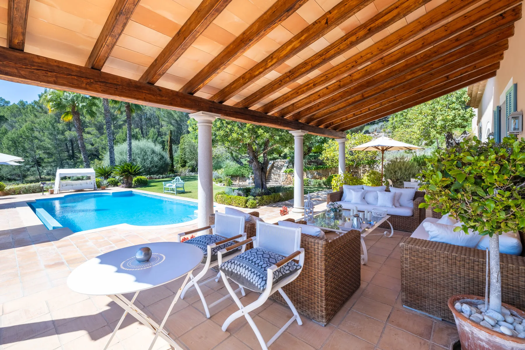 A covered patio with outdoor seating overlooks a swimming pool and garden.