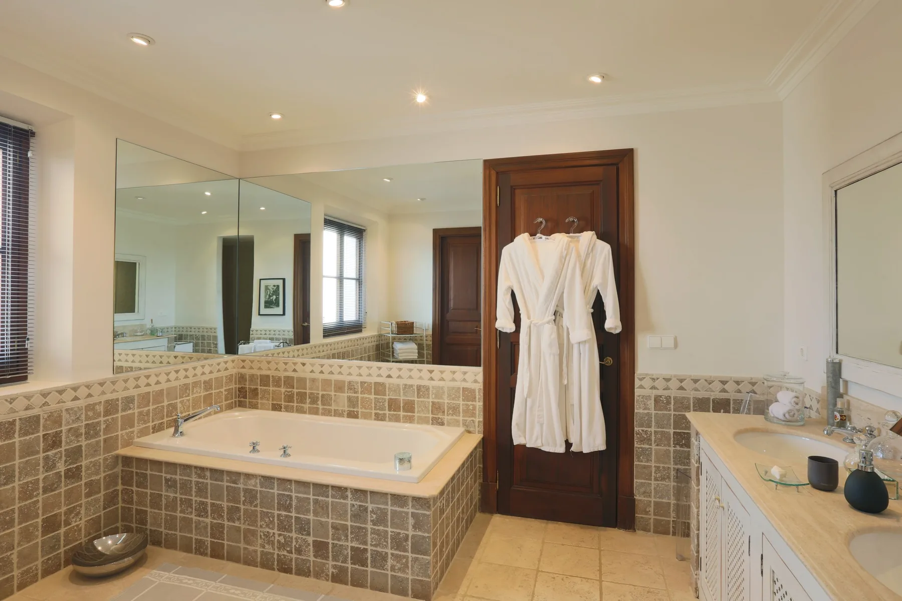 A spacious bathroom features a tiled bathtub, mirrored wall, and white robes hanging on a door.