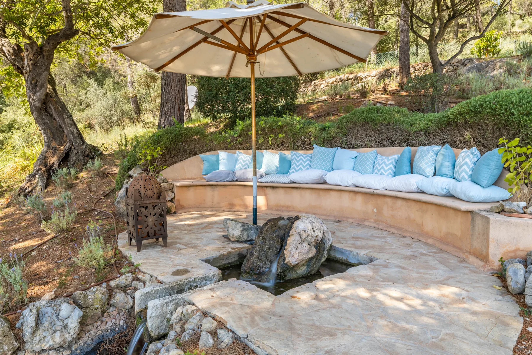 An outdoor stone seating area sits beneath a large umbrella among trees.