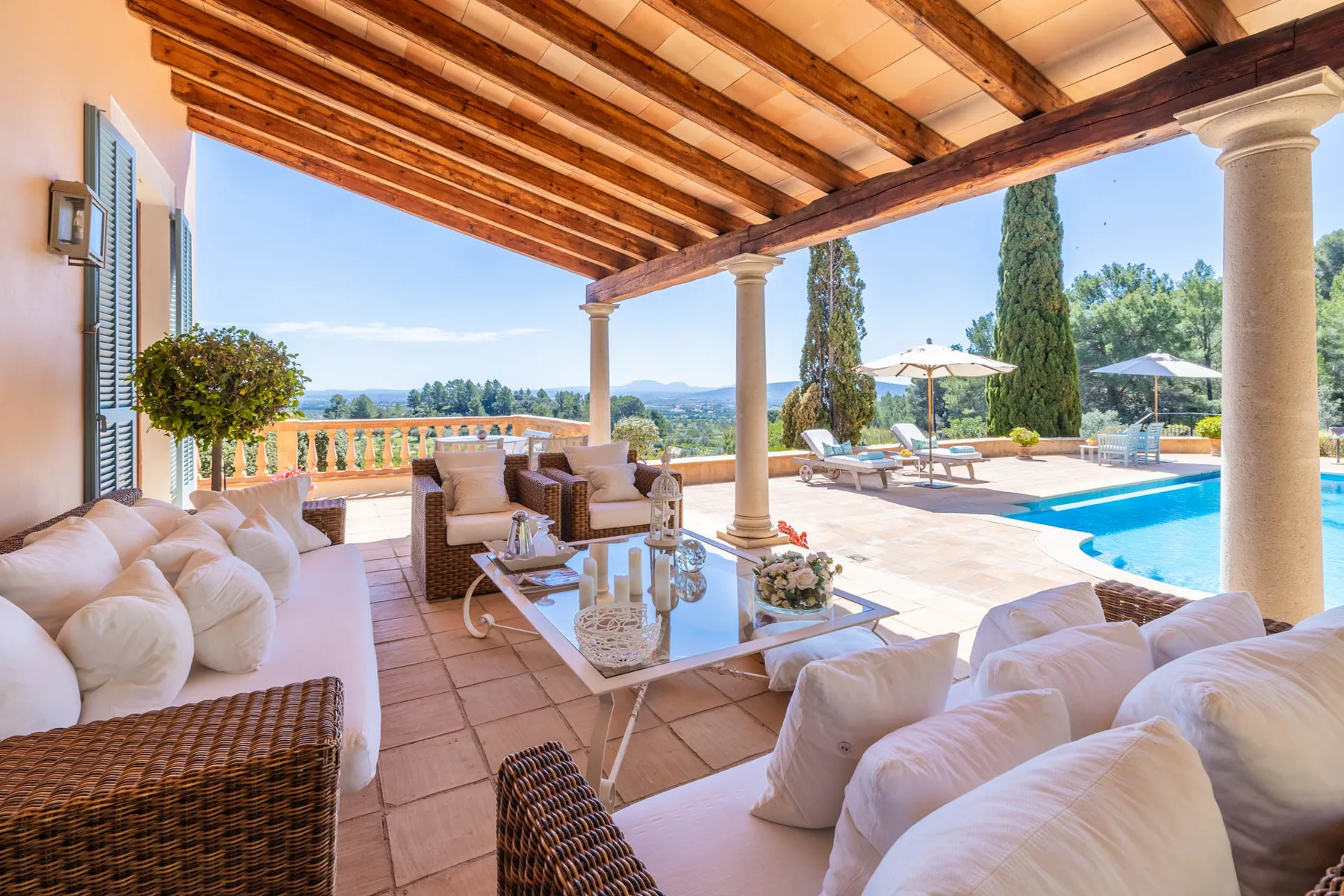 A covered patio with cushioned seating overlooks a swimming pool and distant hills.