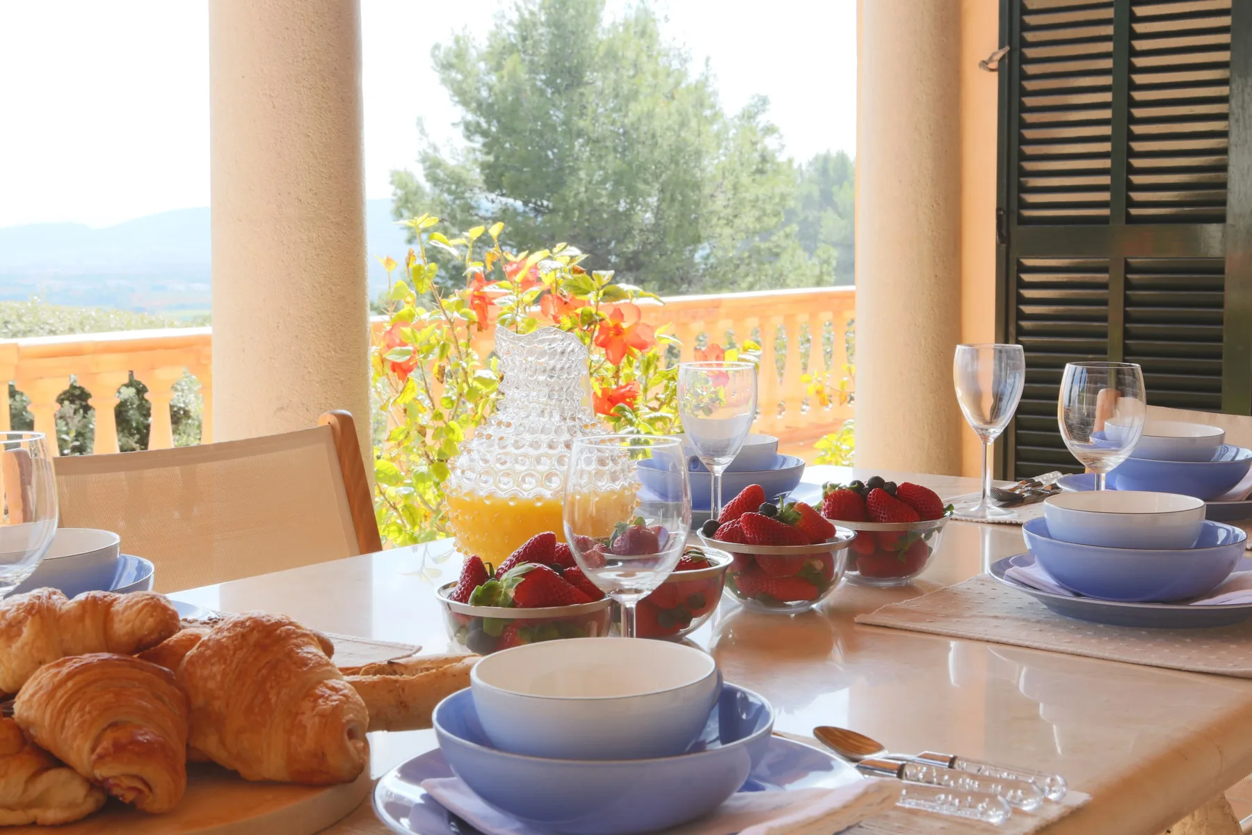 A dining table is set with bowls, glasses, and breakfast pastries beside an open terrace.