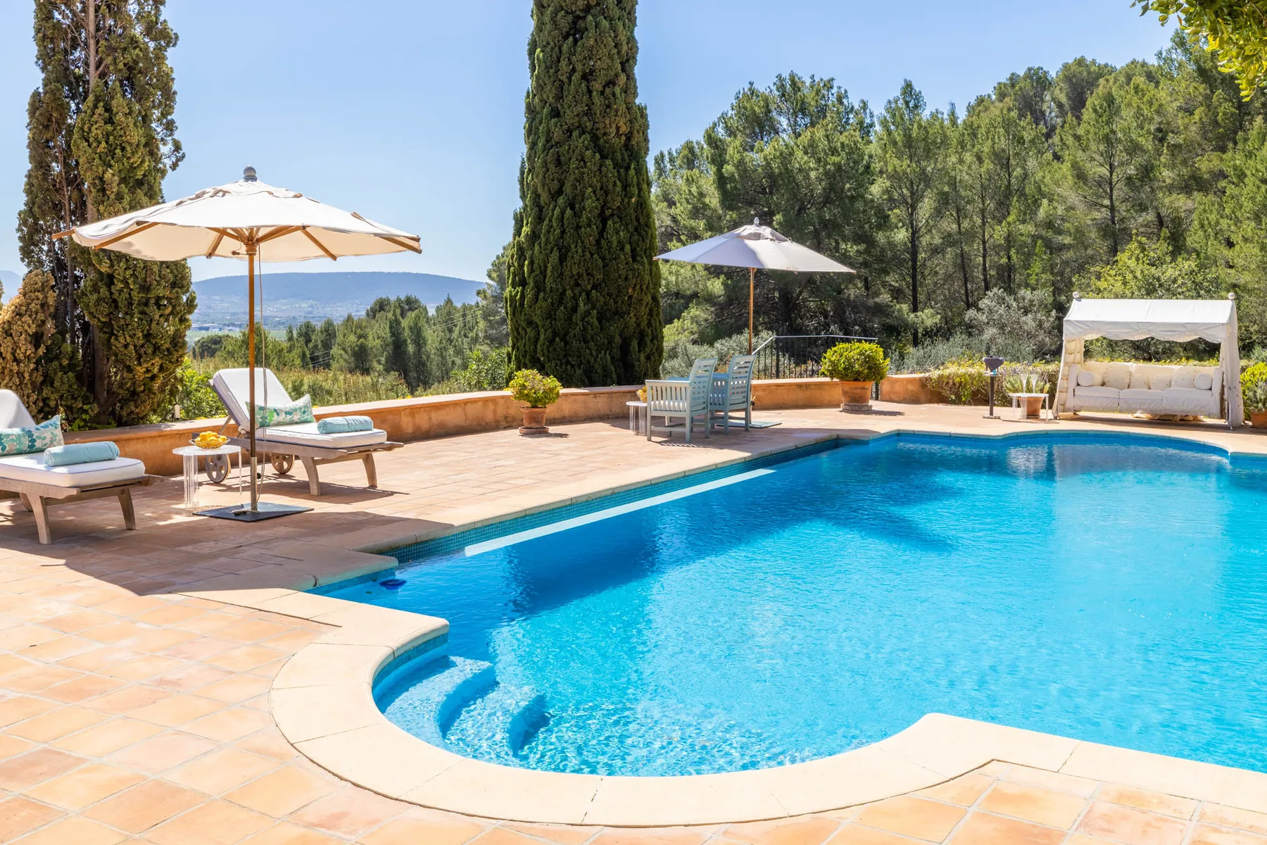 An outdoor swimming pool with a tiled patio, umbrellas, and surrounding trees.