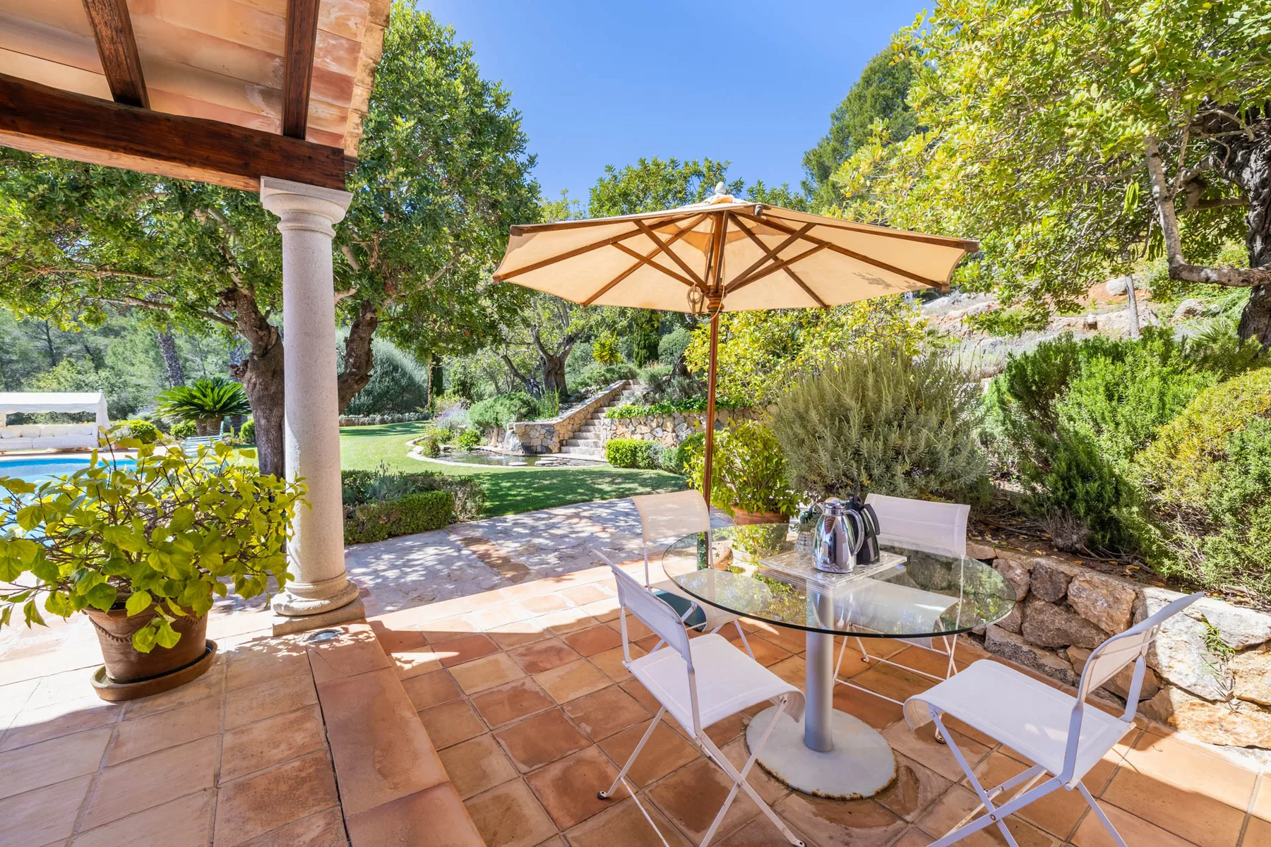 A sunny patio has a table and seating beneath a large umbrella, surrounded by greenery.