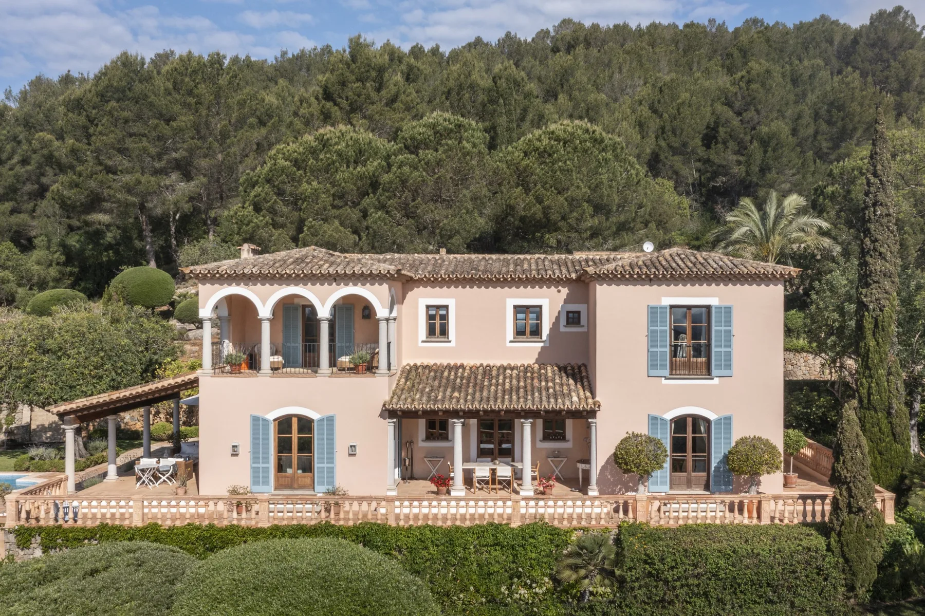 A pink two-story house with tiled roofs and a wooded hillside behind it.