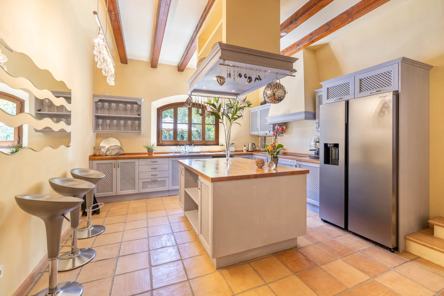 A bright kitchen with a central island, tiled floor, and exposed ceiling beams.