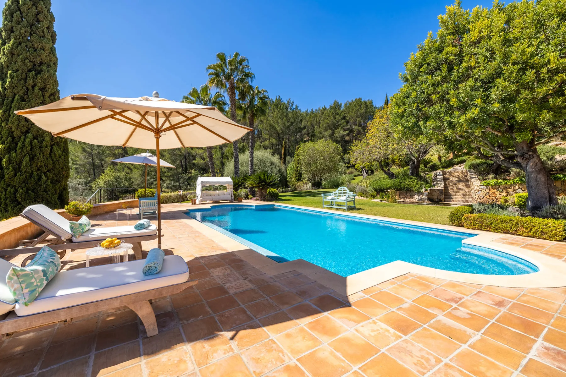 An outdoor swimming pool is surrounded by a tiled patio, loungers, and greenery.