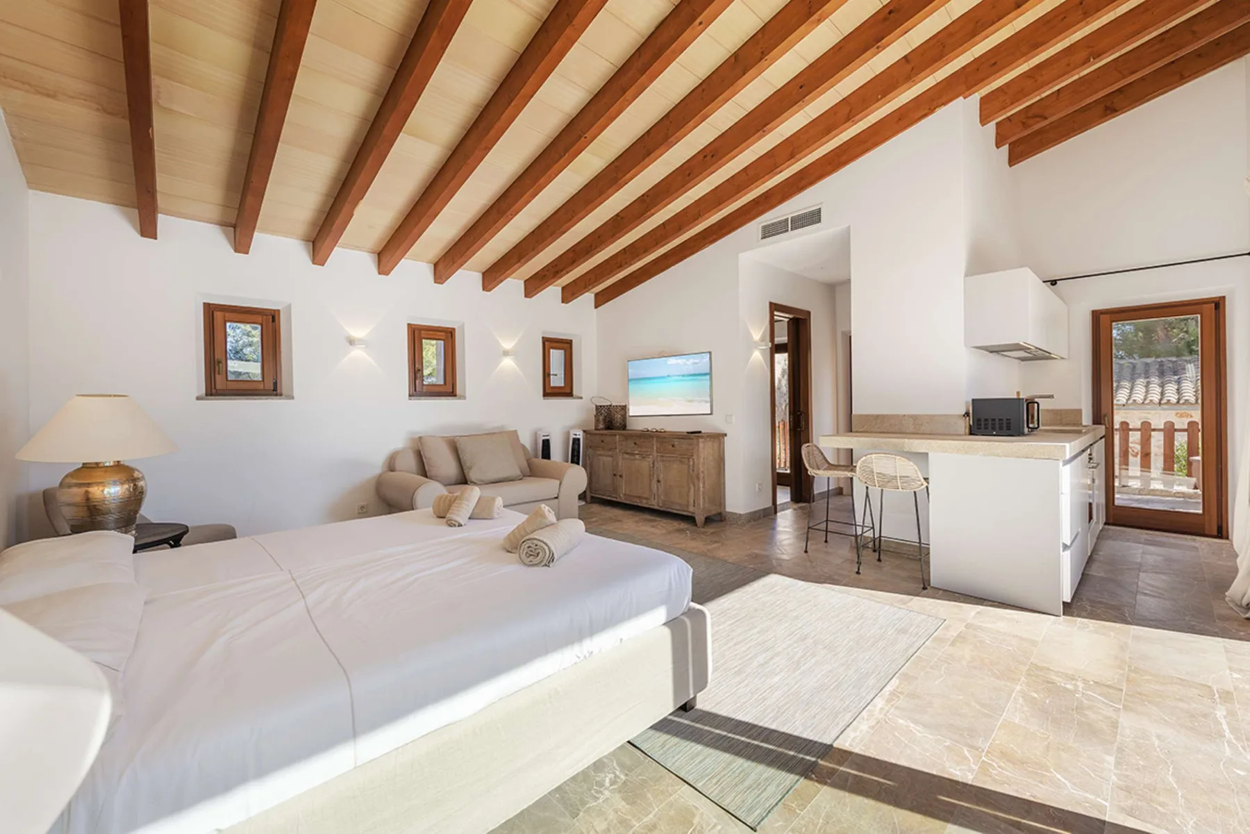A bright bedroom opens onto a compact kitchen, with exposed wooden ceiling beams.
