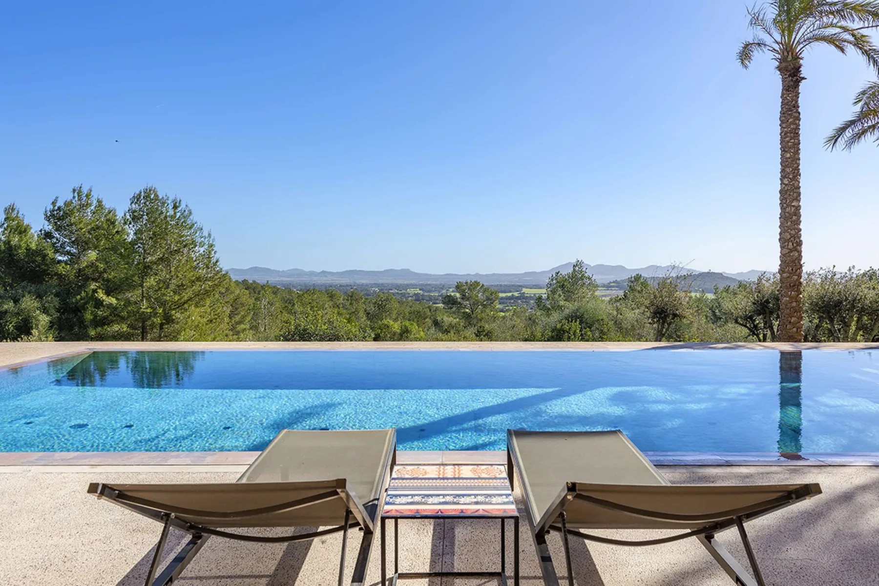 An outdoor swimming pool overlooks a tree-lined landscape, with two loungers in the foreground.