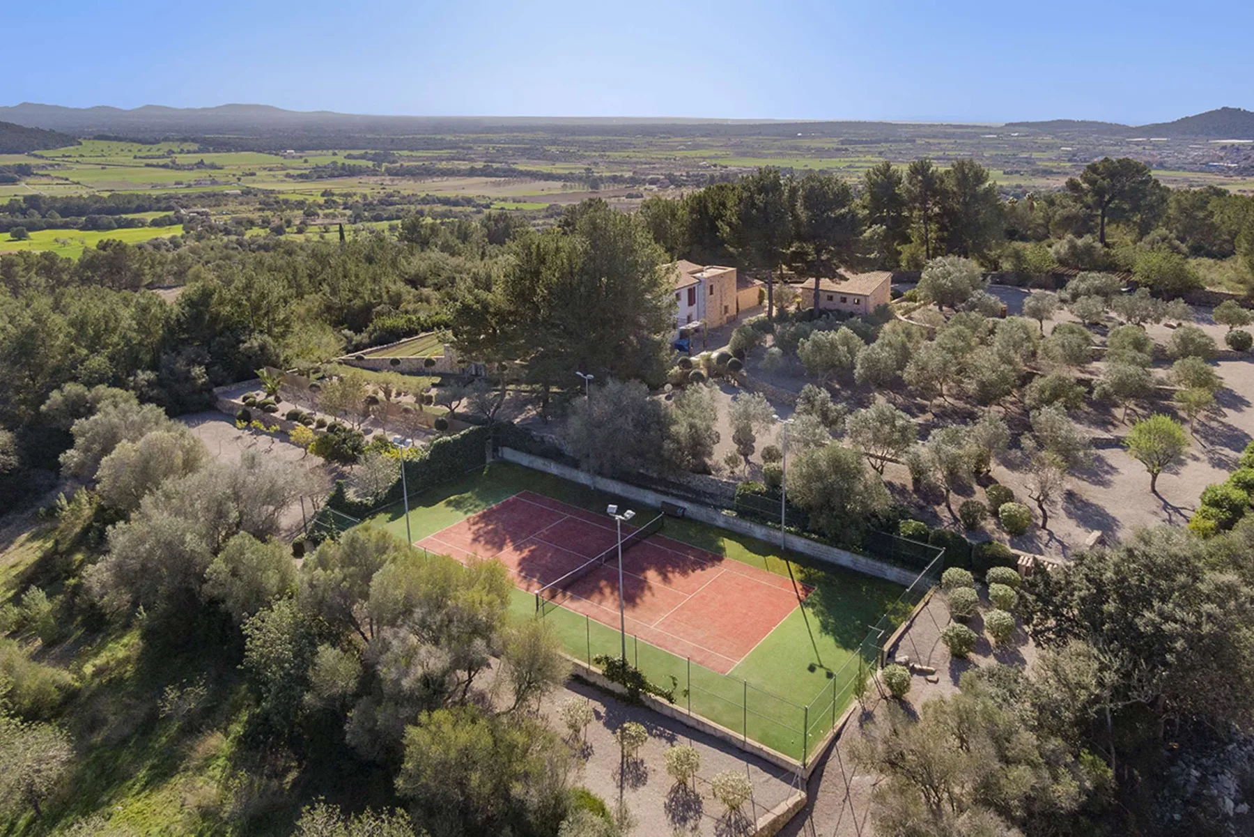 An aerial view shows a tennis court beside a house amid wooded countryside.