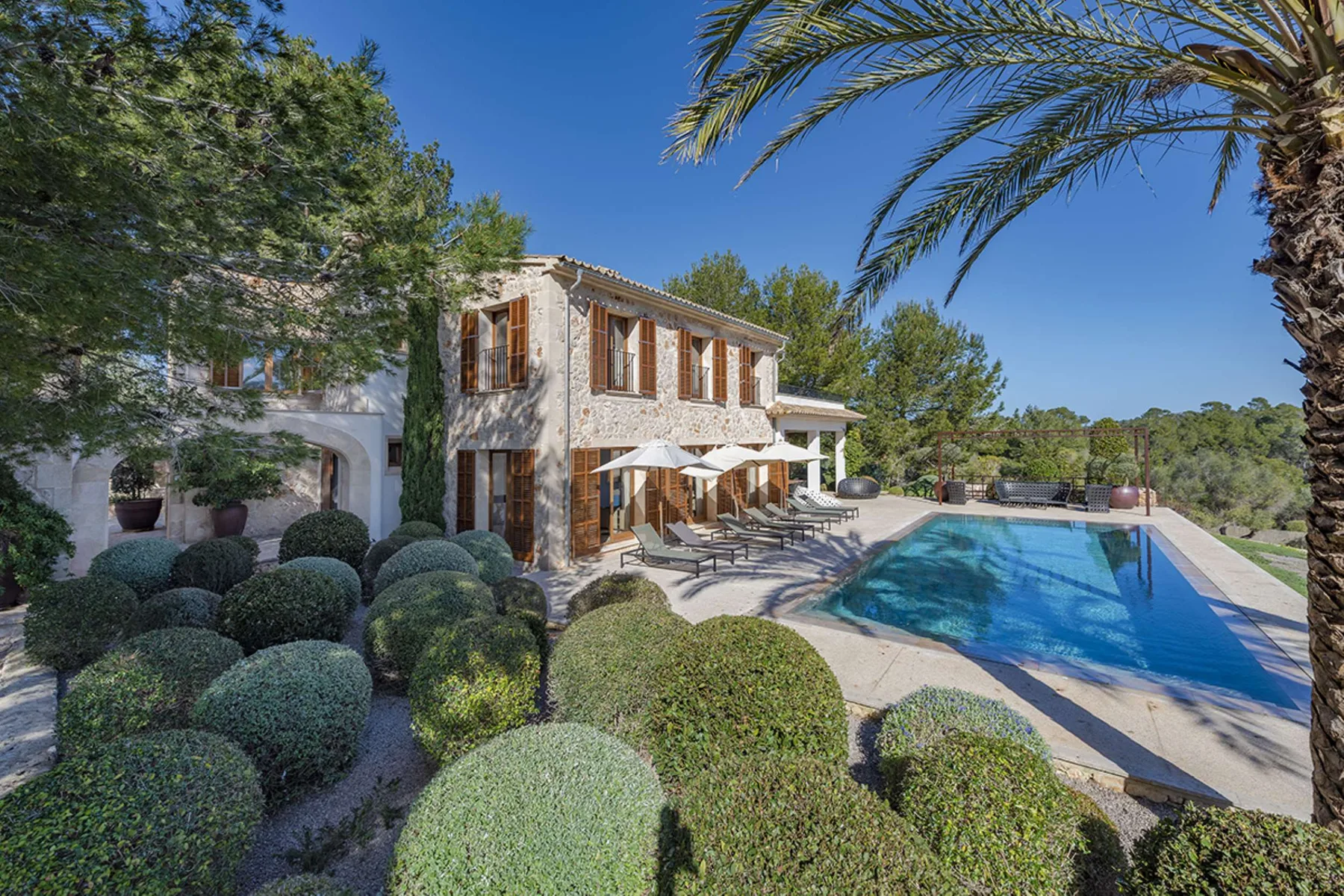 A two-story villa overlooks a swimming pool, with trimmed shrubs in the foreground.