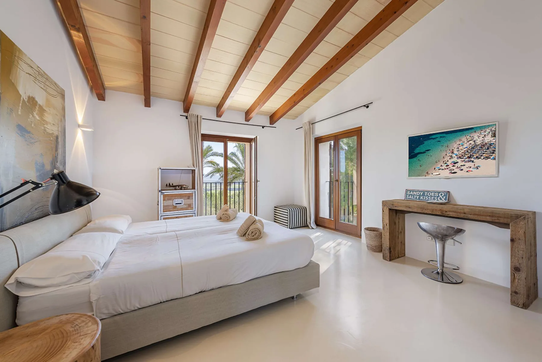 A bright bedroom with a large bed beneath exposed wooden ceiling beams.