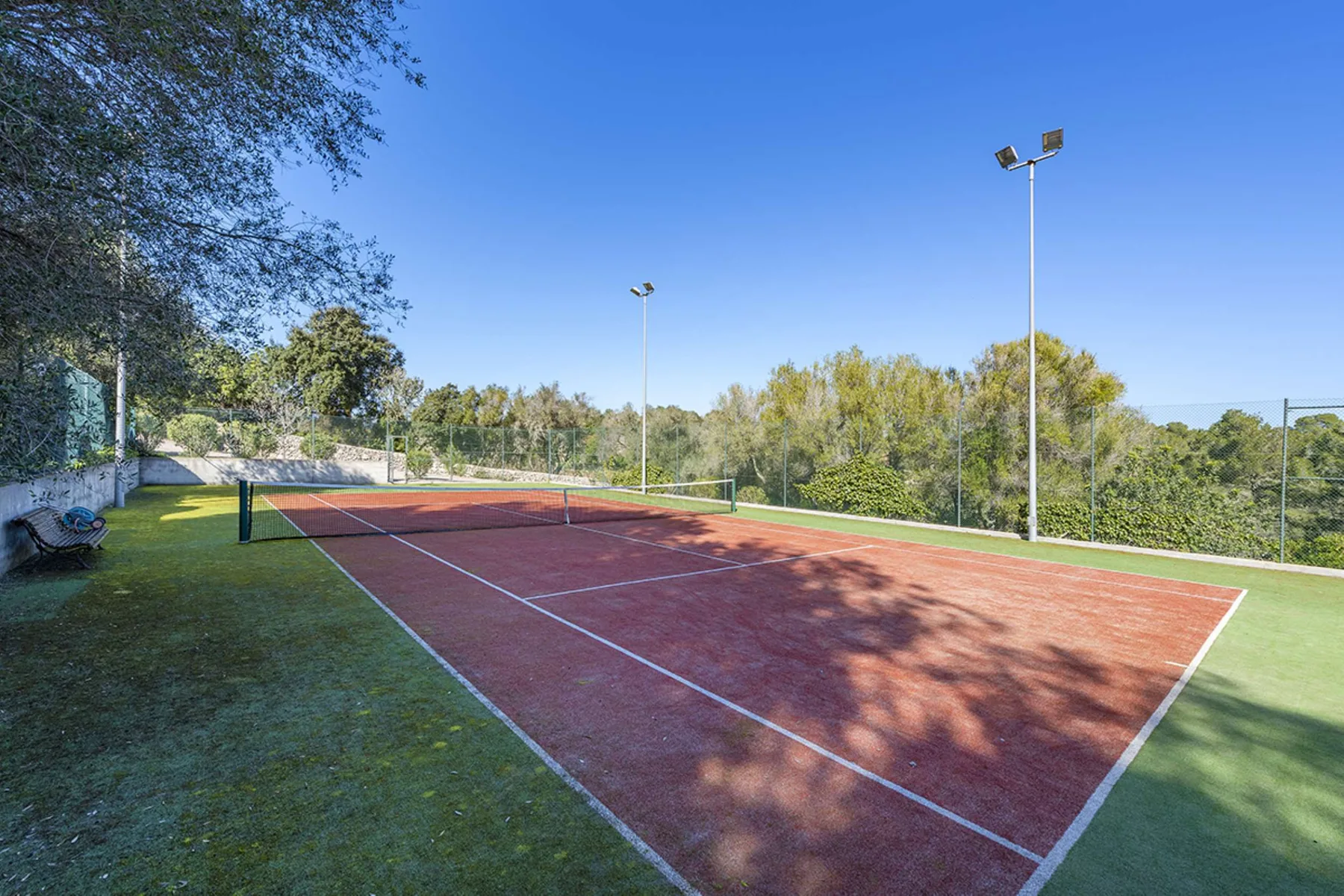 An outdoor tennis court with a net, surrounded by trees and fencing.