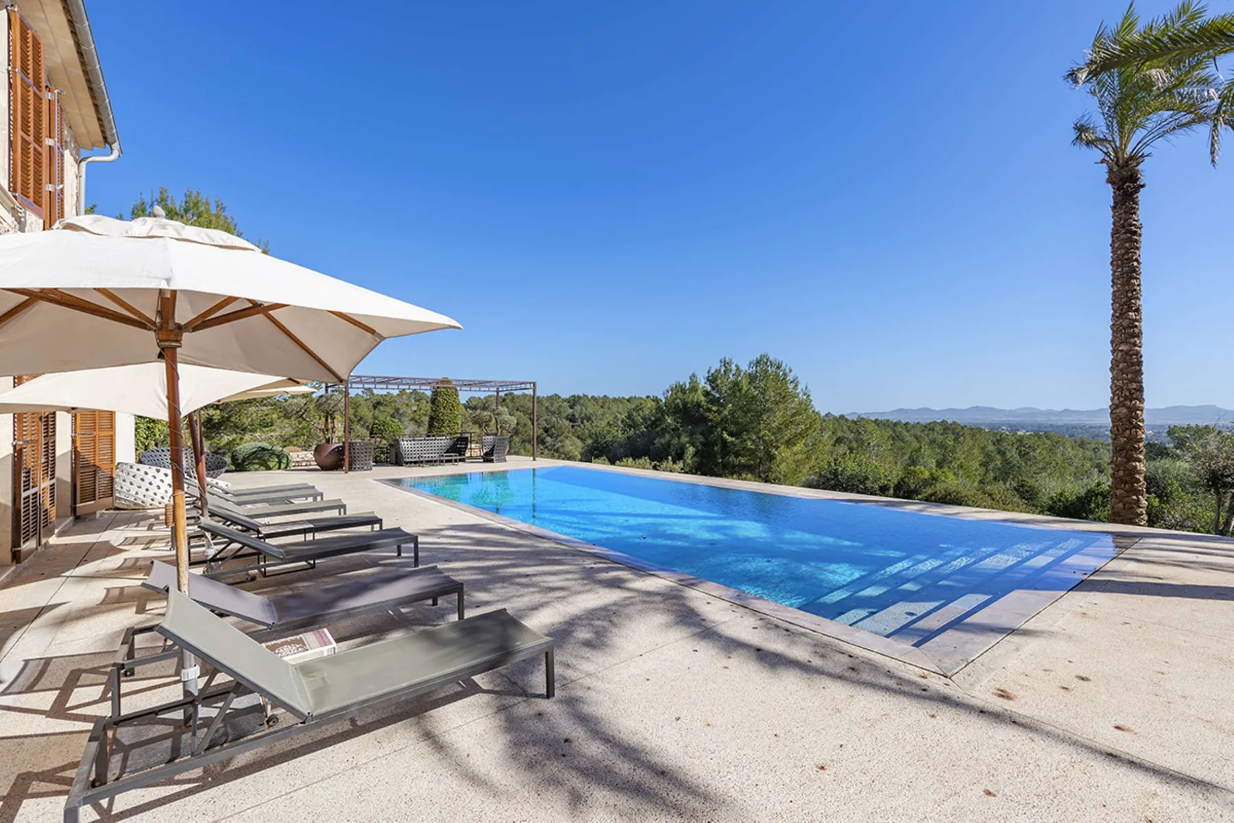 An outdoor swimming pool beside a sunny patio with lounge seating and an umbrella.