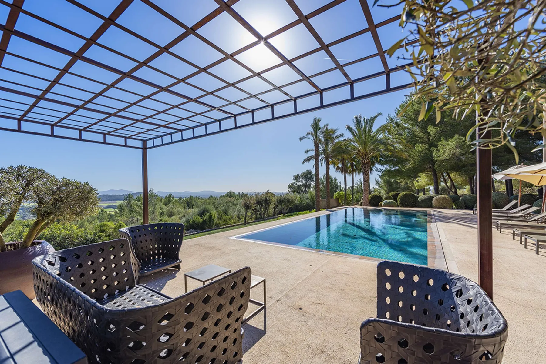 An outdoor pool and patio seating beneath a pergola overlook a green landscape.