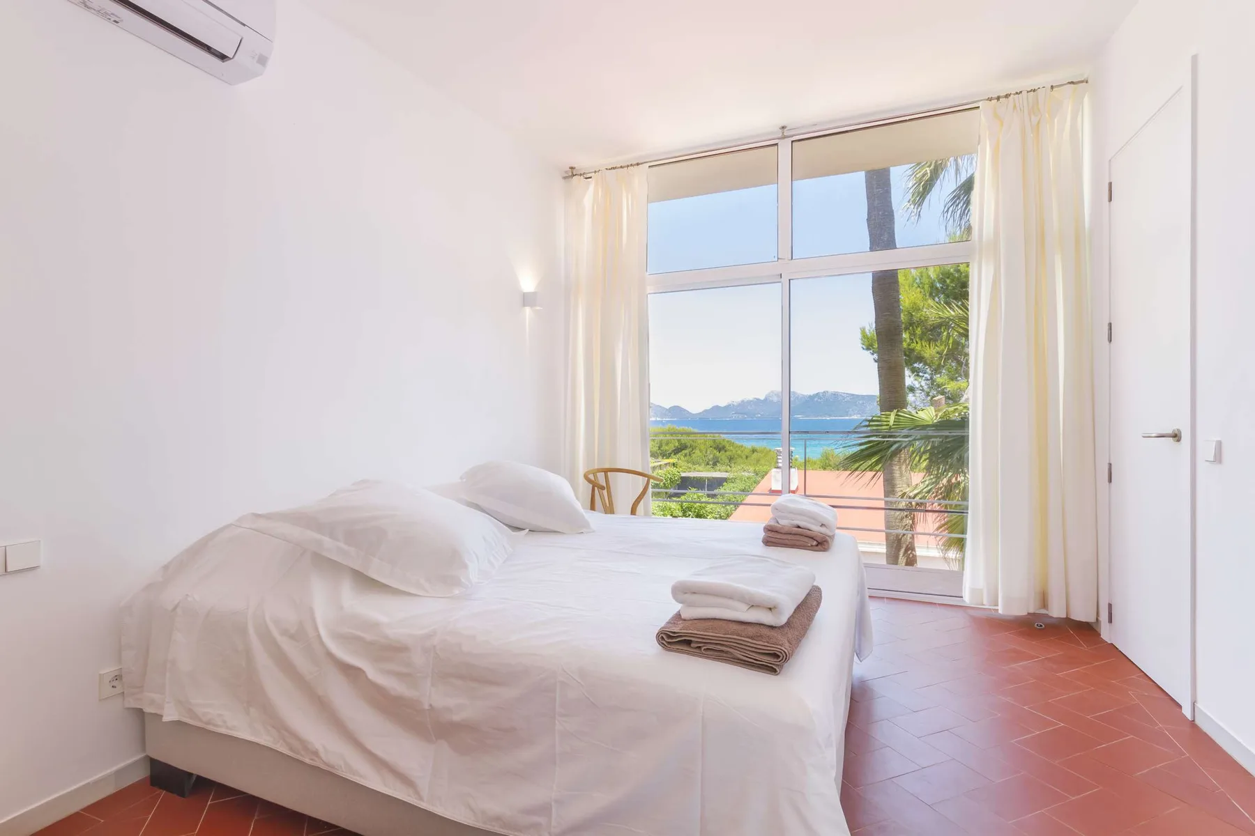 A bright bedroom with a bed beside a large window overlooking the sea.