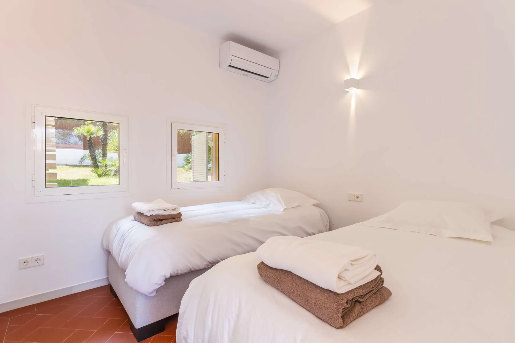 A bright bedroom with two beds, windows, and a wall-mounted air conditioner.