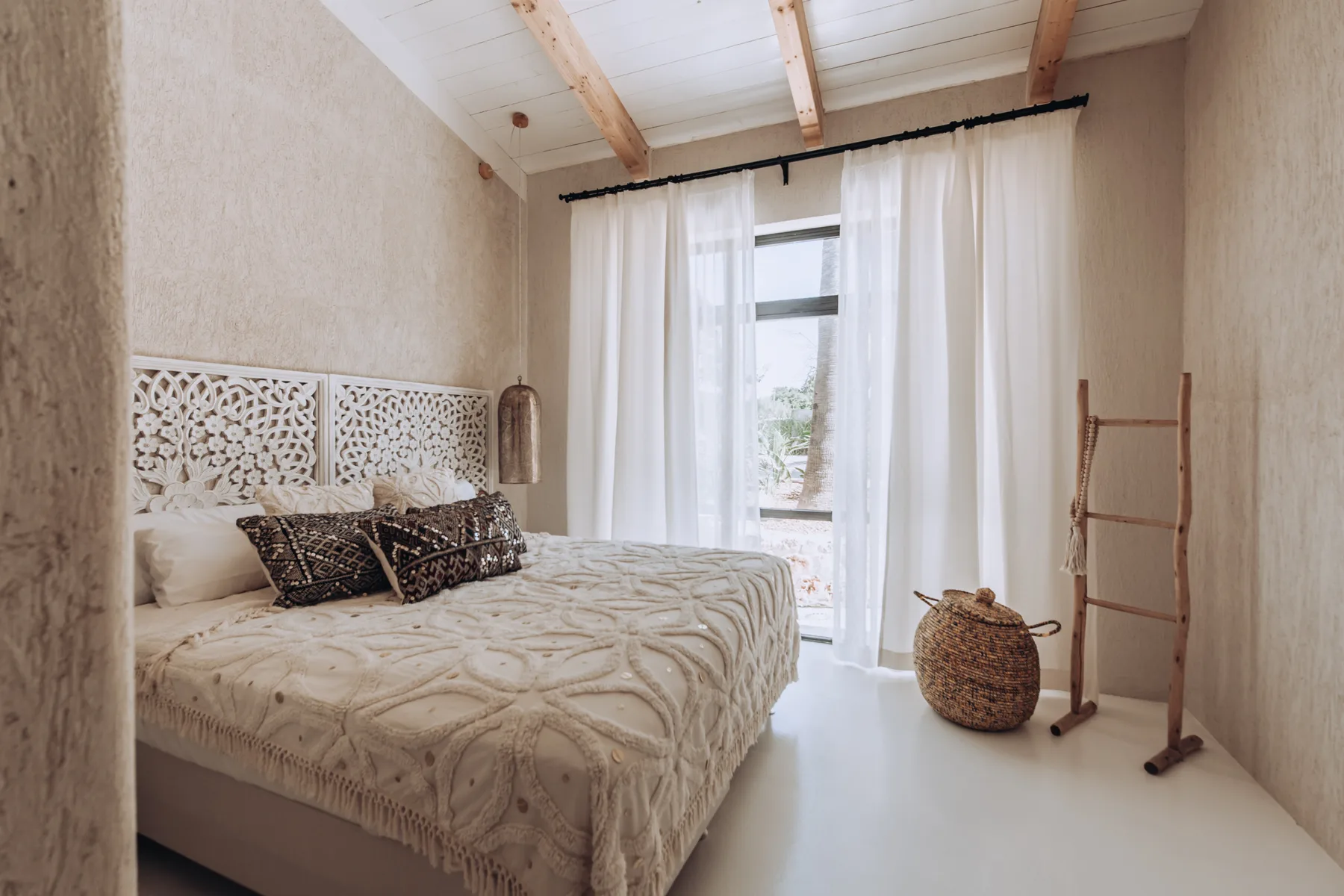 A bedroom with a bed beside sheer curtains and a bright window.