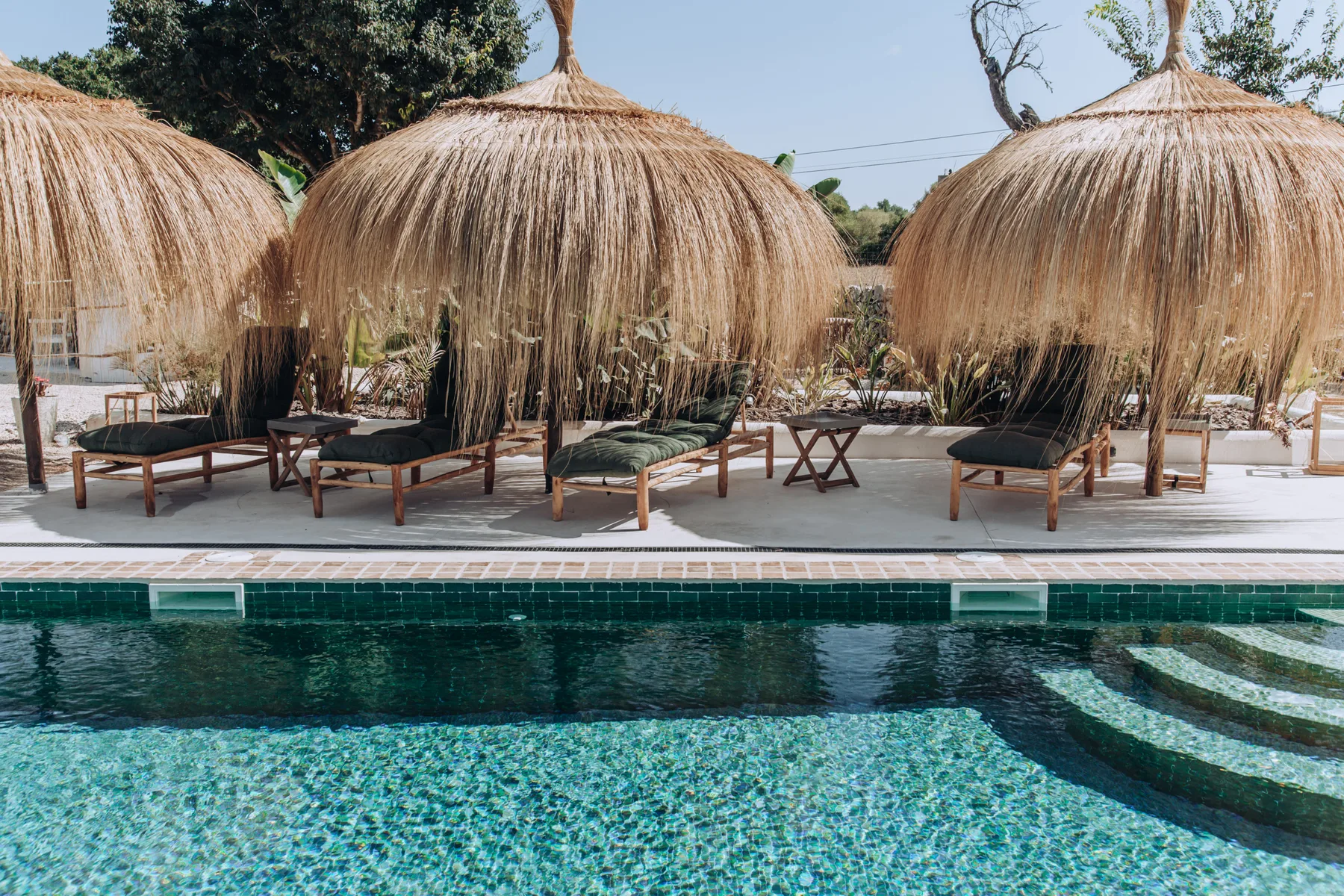 A swimming pool sits beside shaded lounge seating beneath thatched umbrellas.