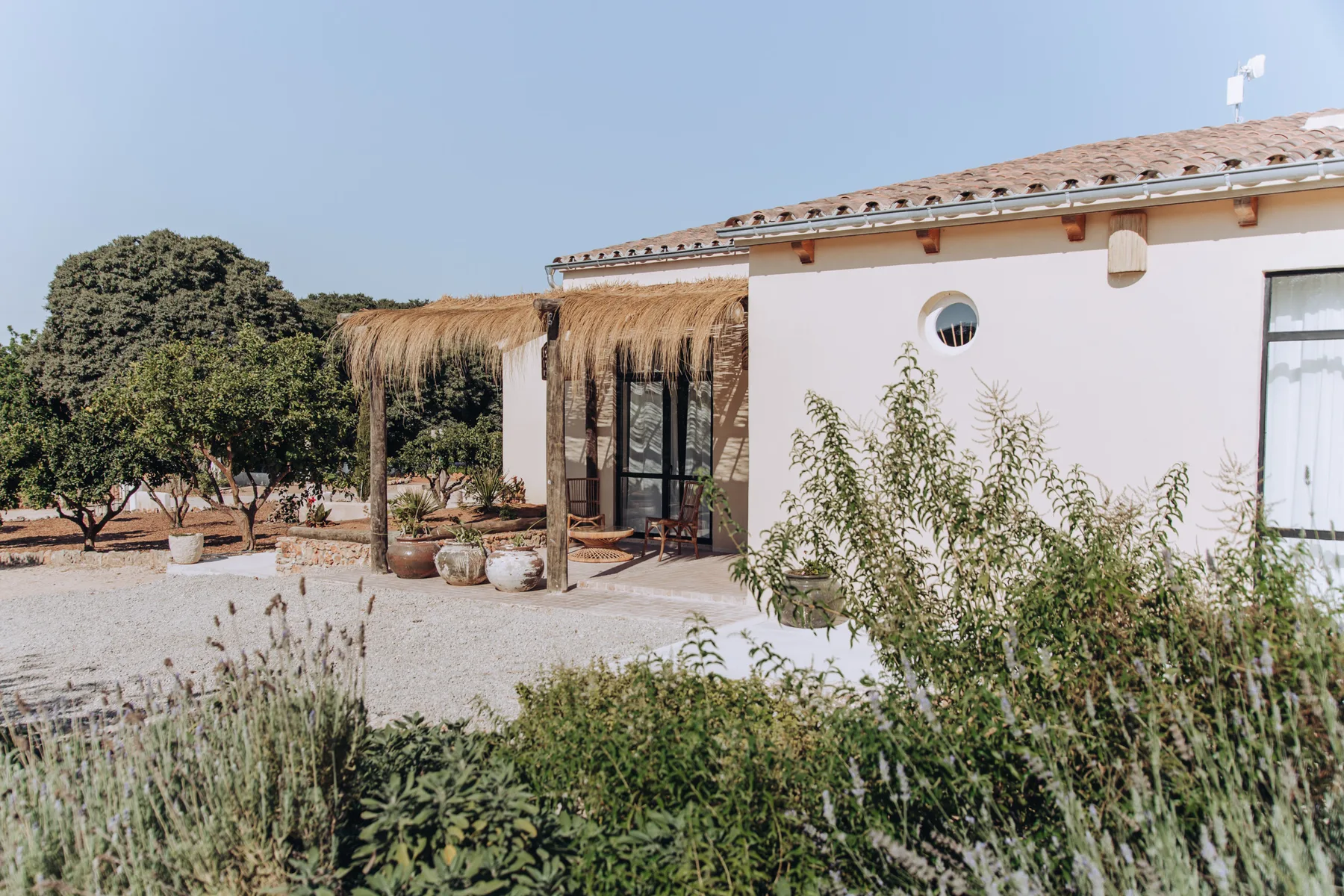A white building opens onto a gravel courtyard with a thatched shade structure.