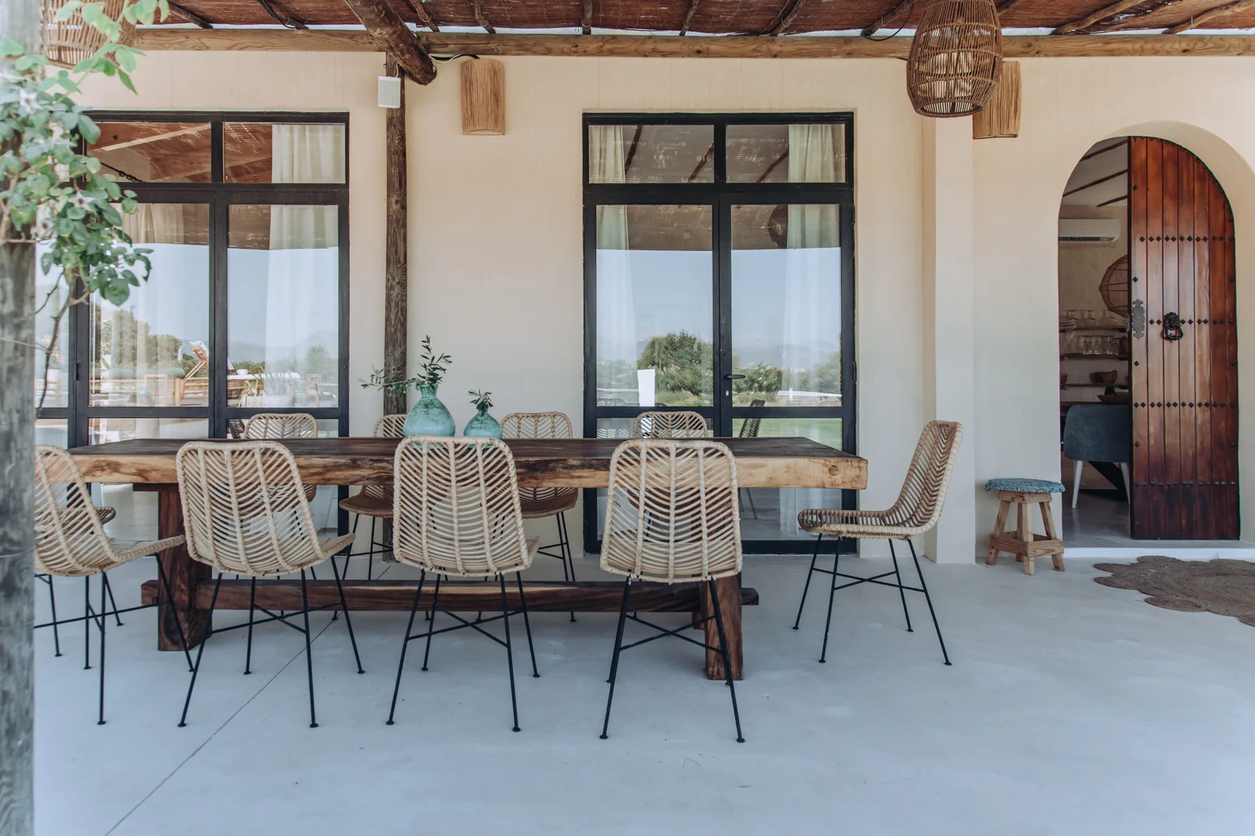A dining area with a long table and woven seating beneath a shaded roof.
