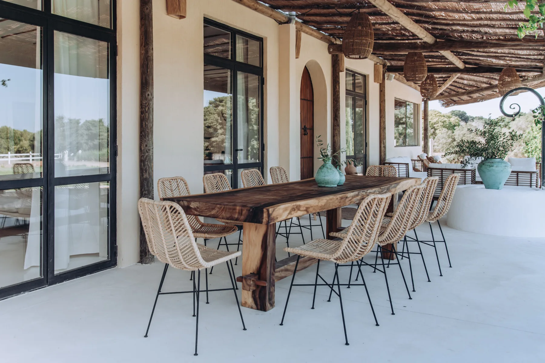 A covered outdoor dining area with a long wooden table and woven seating.