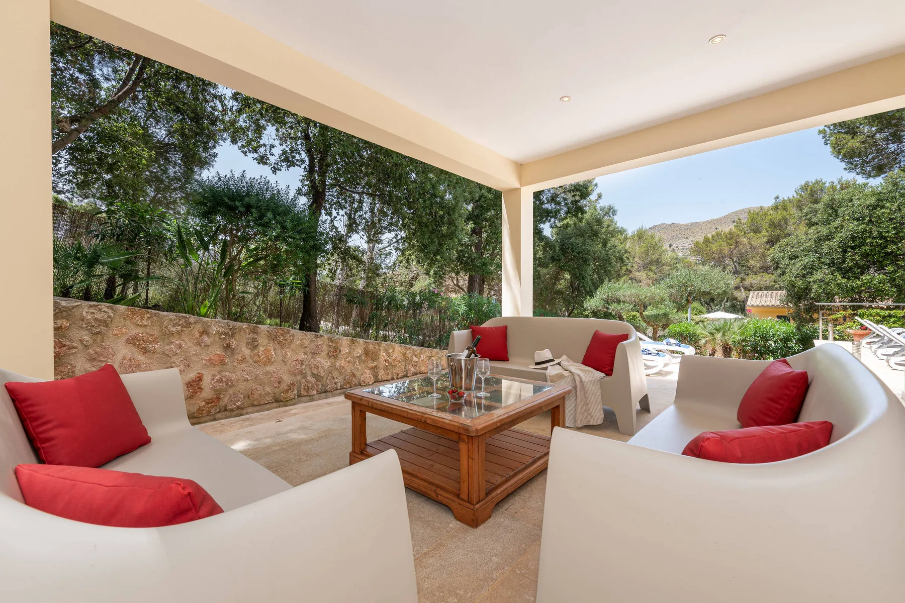 A covered outdoor seating area with red cushions and a glass-topped table overlooks a garden.