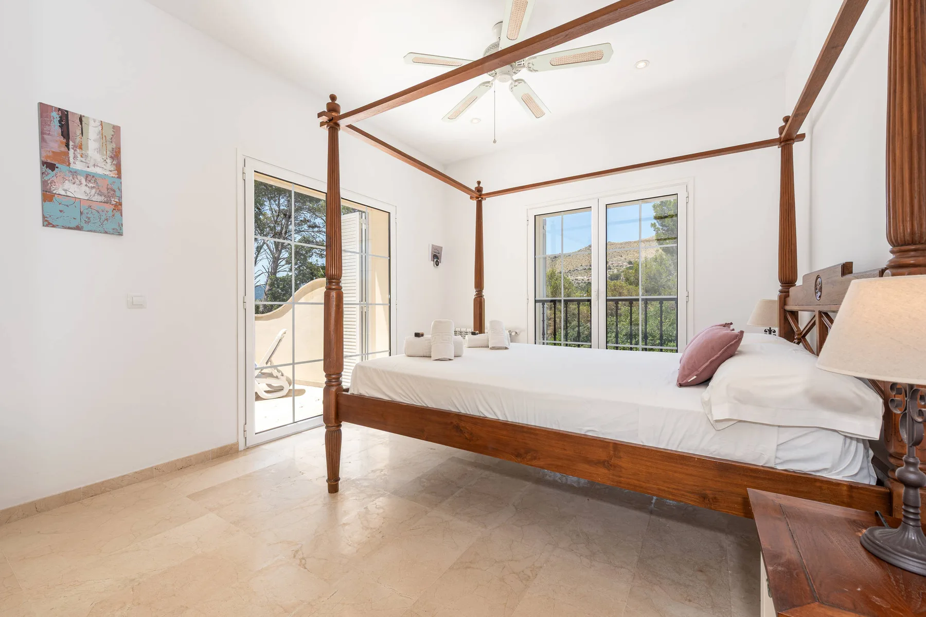 A bright bedroom features a wooden four-poster bed beside glass doors opening to a balcony.