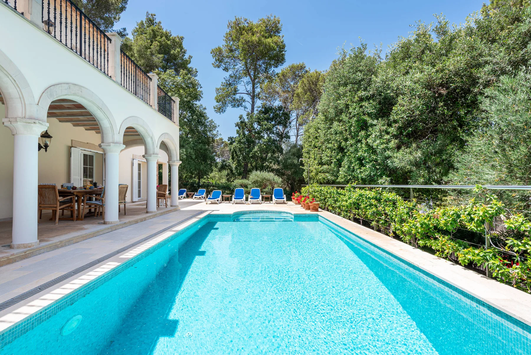 An outdoor swimming pool beside a covered terrace with seating and surrounding greenery.
