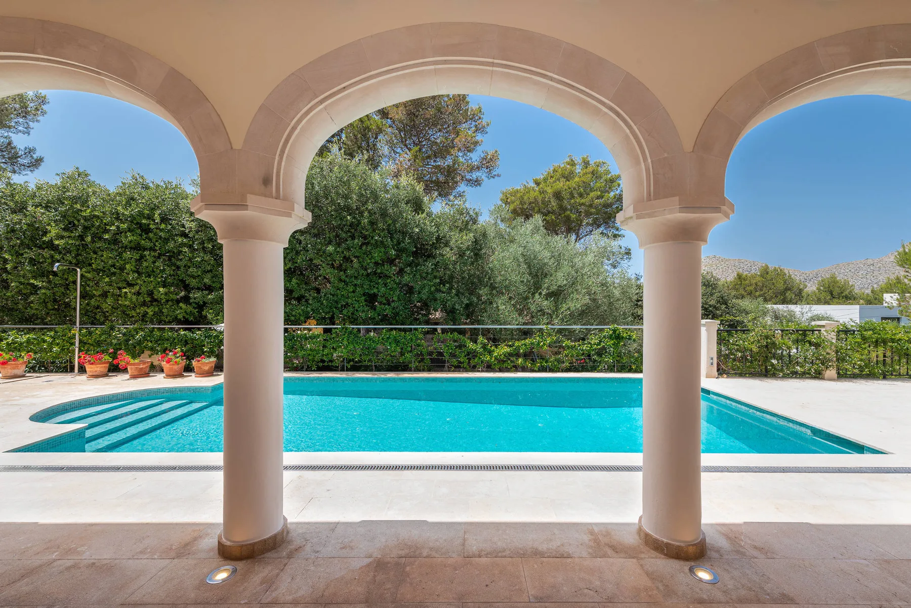 A swimming pool is seen through a row of arched openings, with greenery beyond.