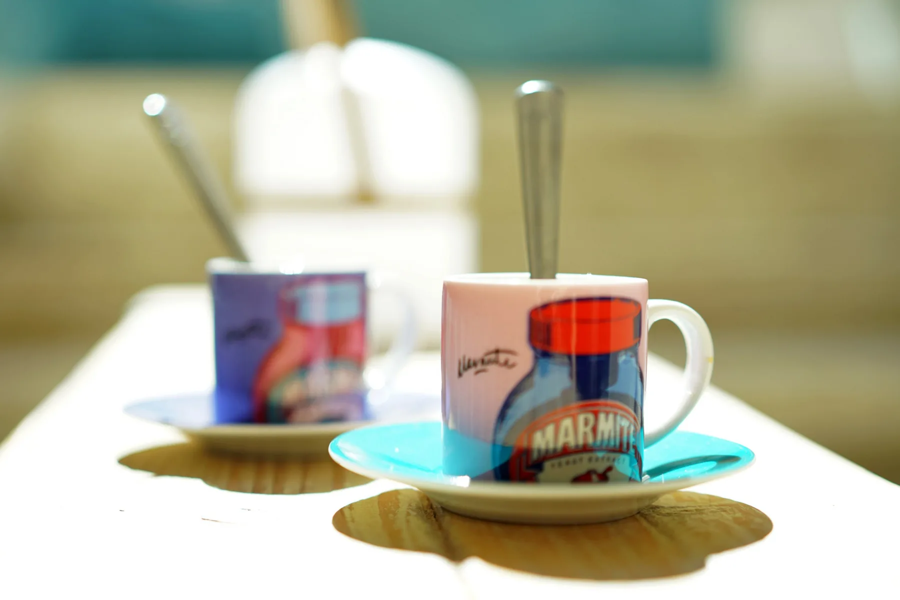 Two mugs with spoons sit on saucers on a bright table.