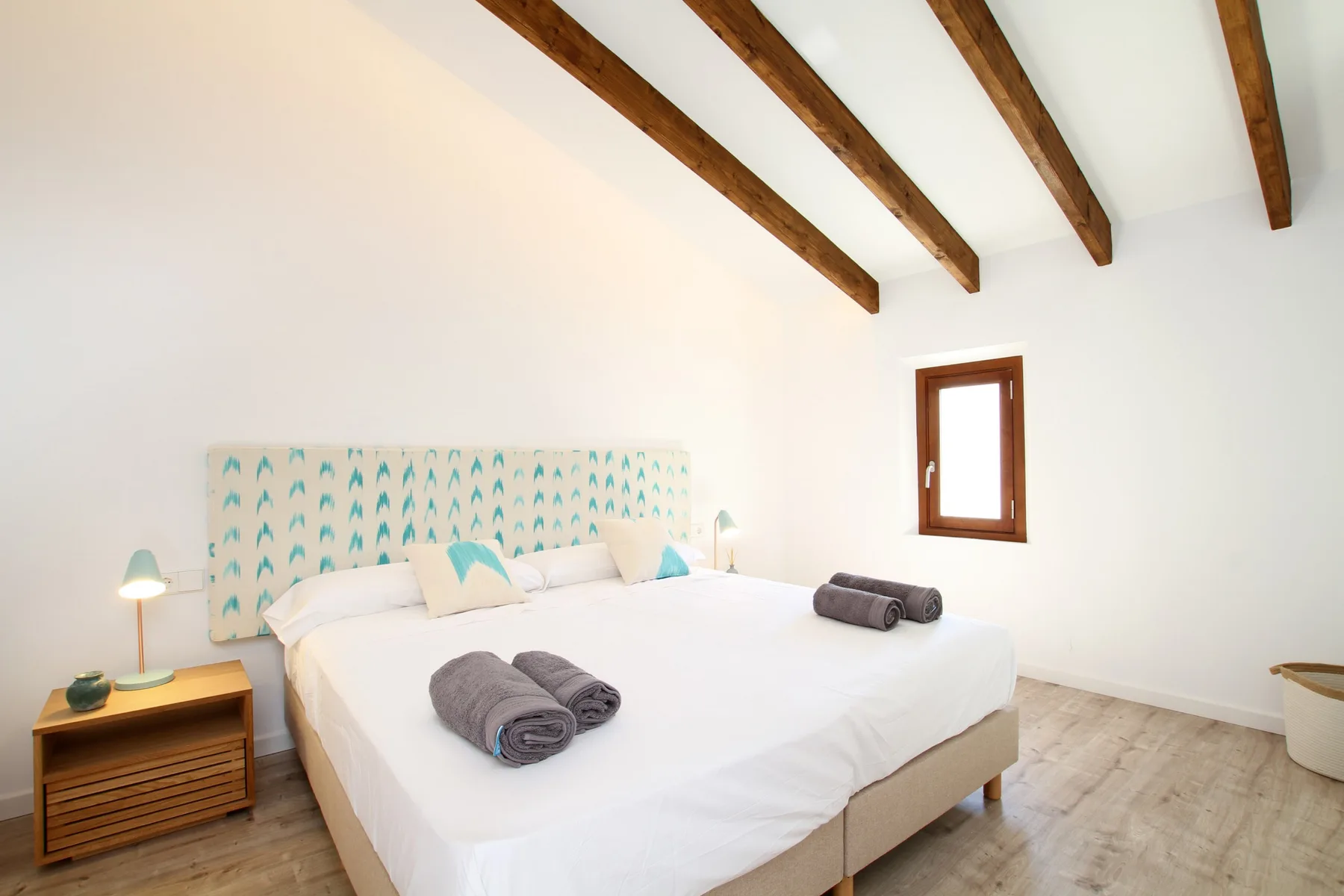 A bright bedroom with a large bed beneath exposed wooden ceiling beams.