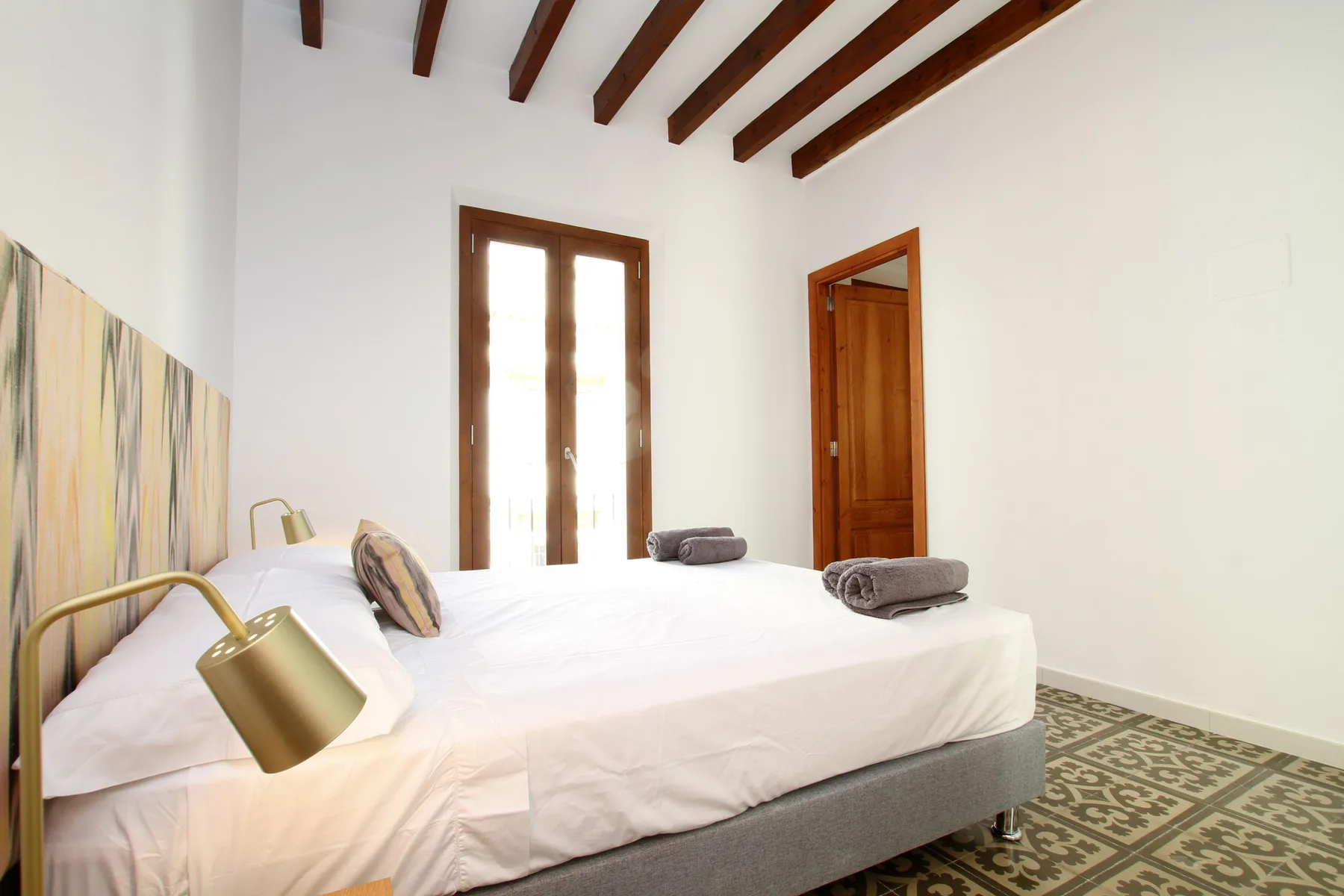 A bright bedroom with a large bed, exposed ceiling beams, and a patterned floor.