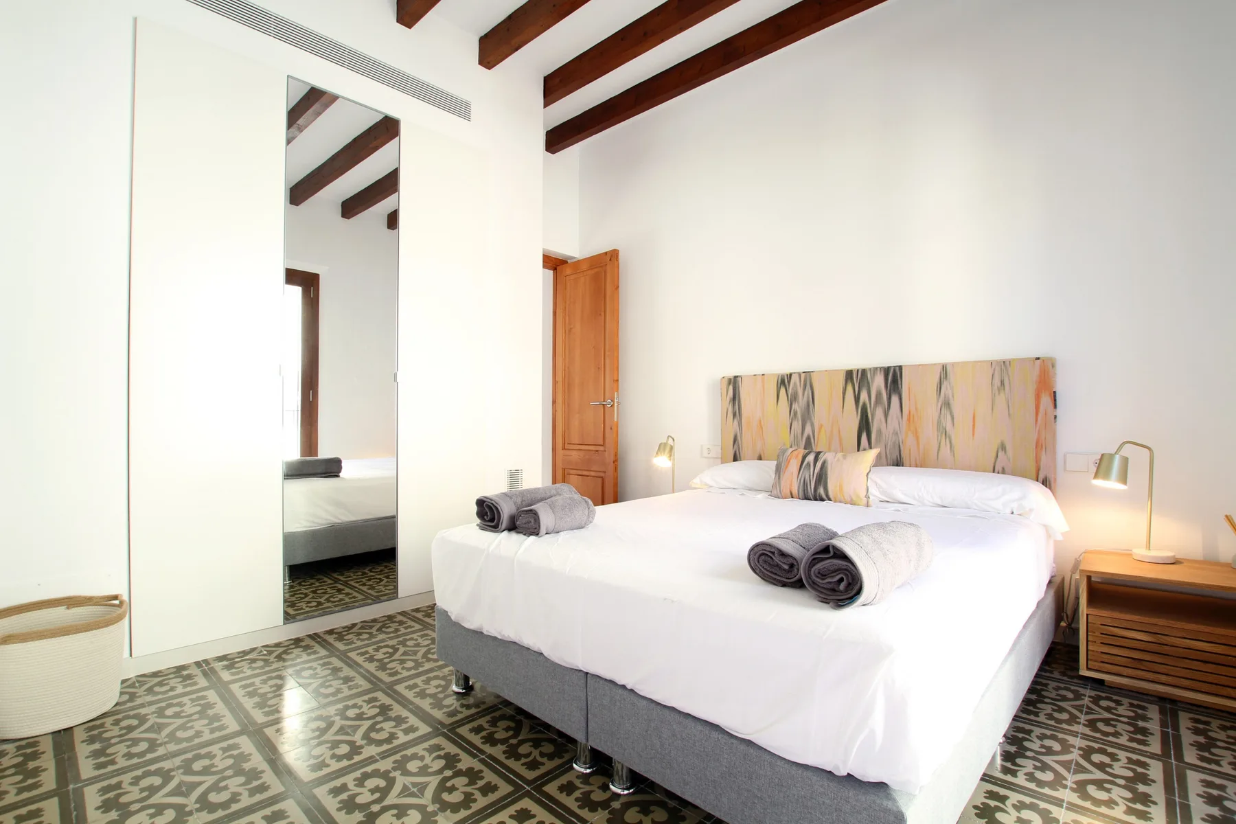 A bright bedroom with a bed, patterned floor tiles, and exposed ceiling beams.