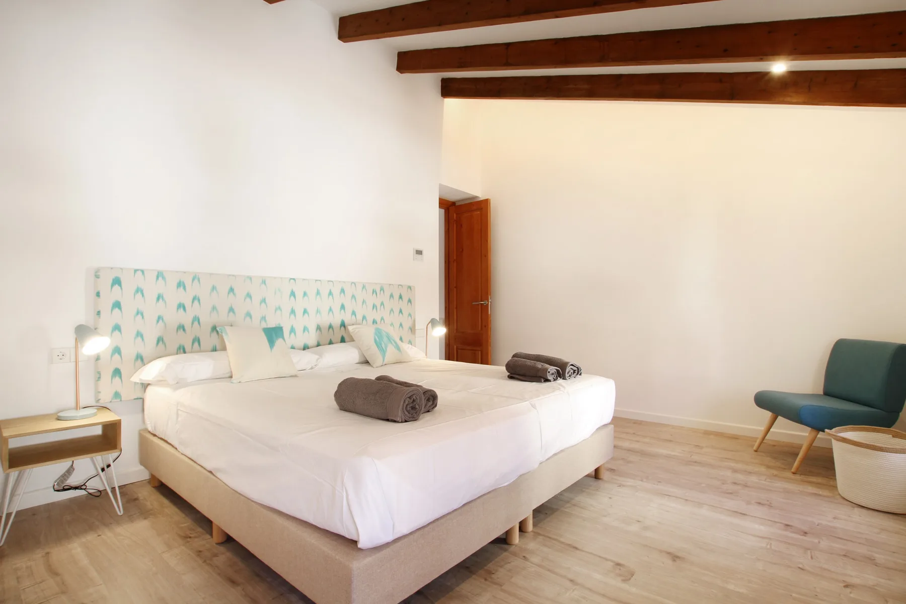 A bright bedroom with a large bed, patterned headboard, and exposed ceiling beams.