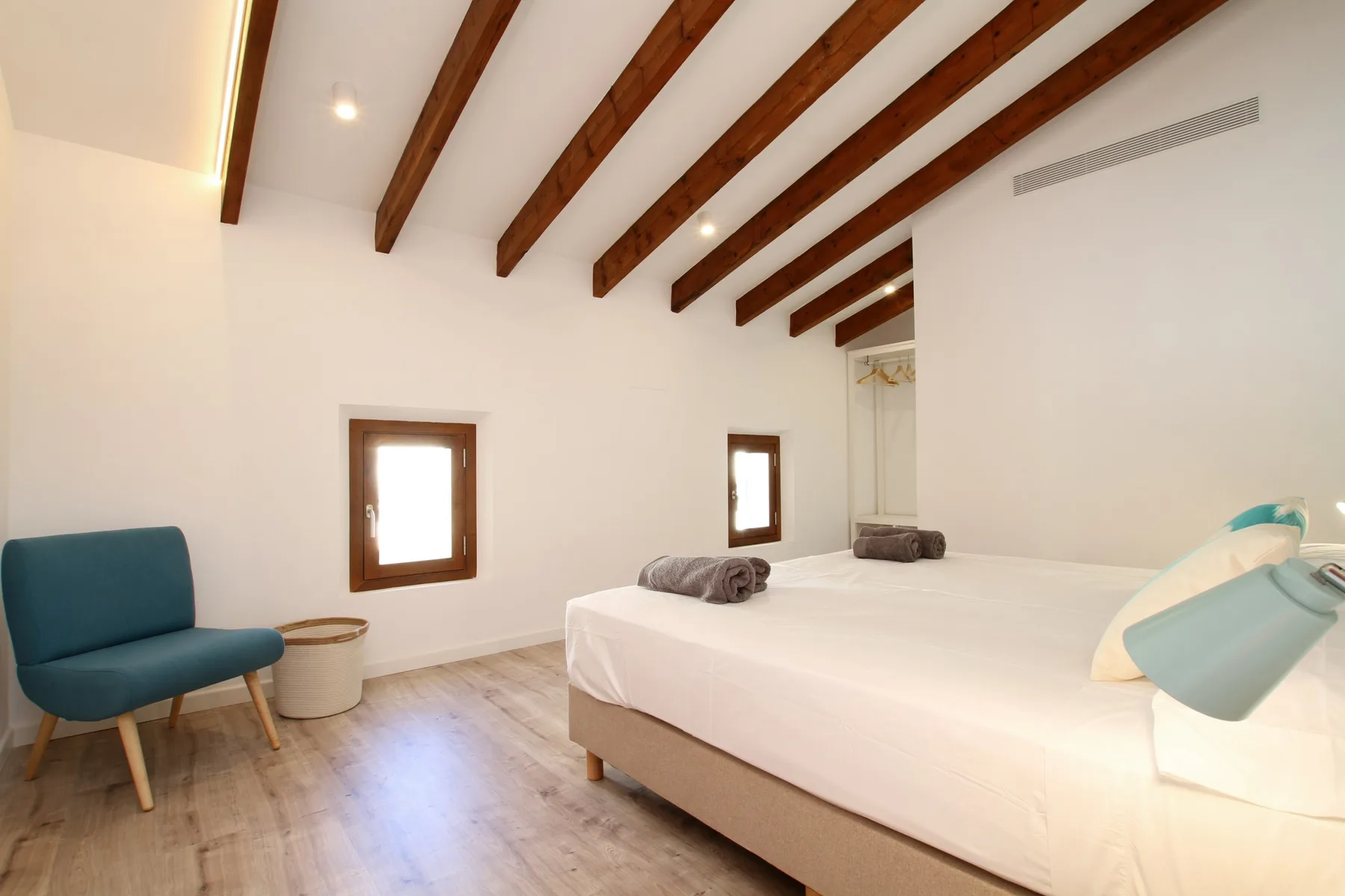 A bright bedroom with a large bed, wooden ceiling beams, and a chair.