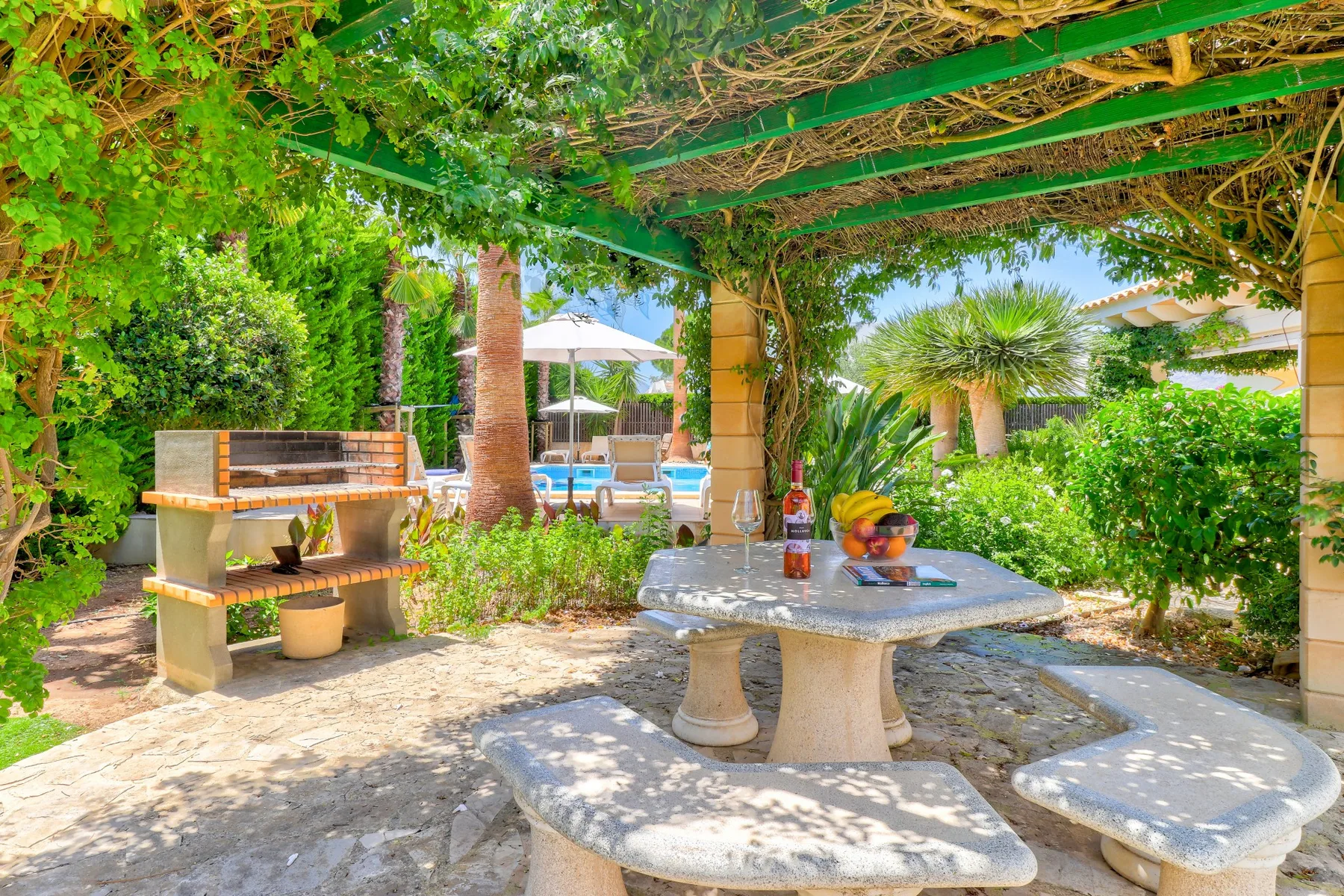 A shaded outdoor dining area beneath a leafy pergola, with a garden and pool beyond.