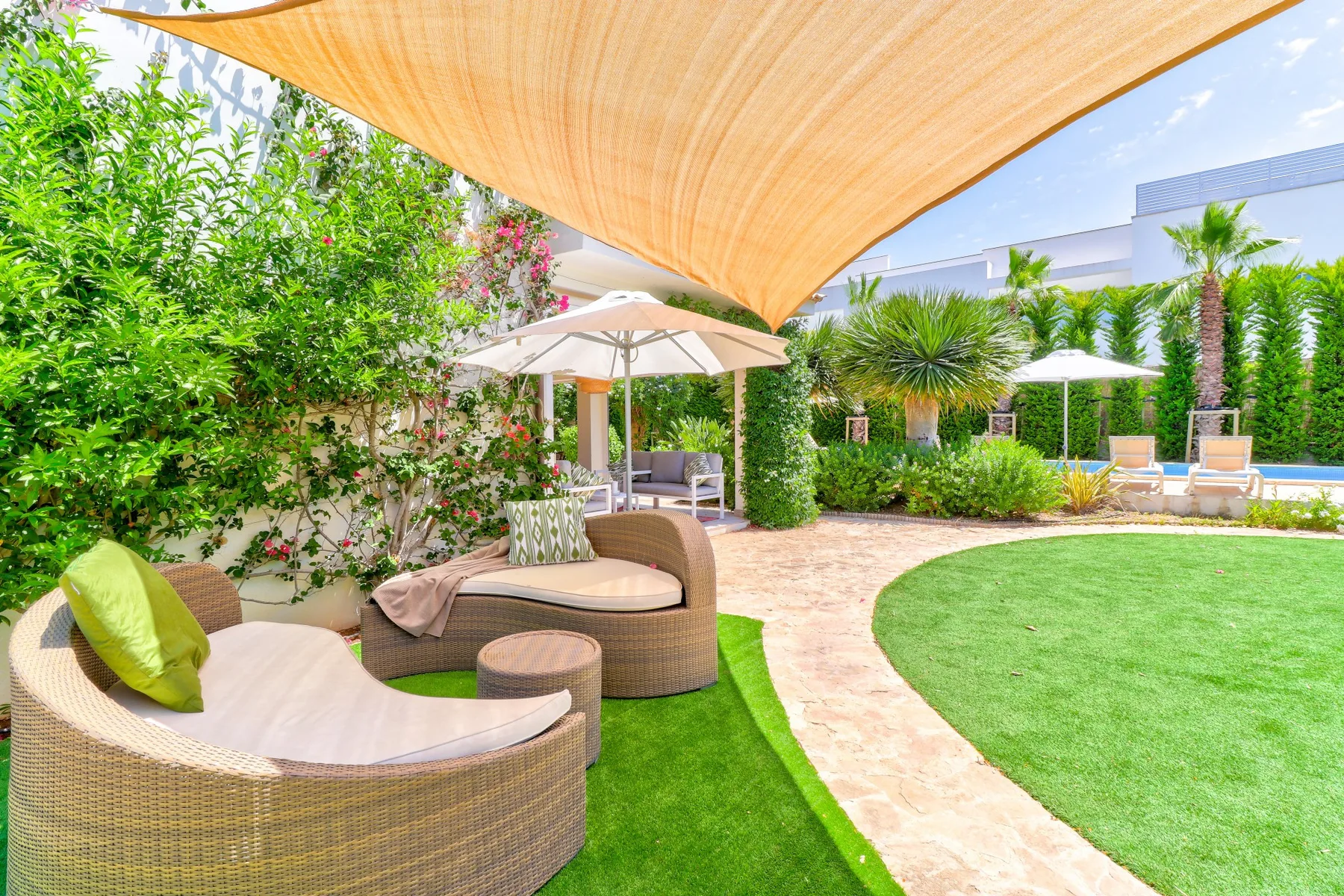 An outdoor patio has curved seating beneath a shade sail, beside a green lawn.