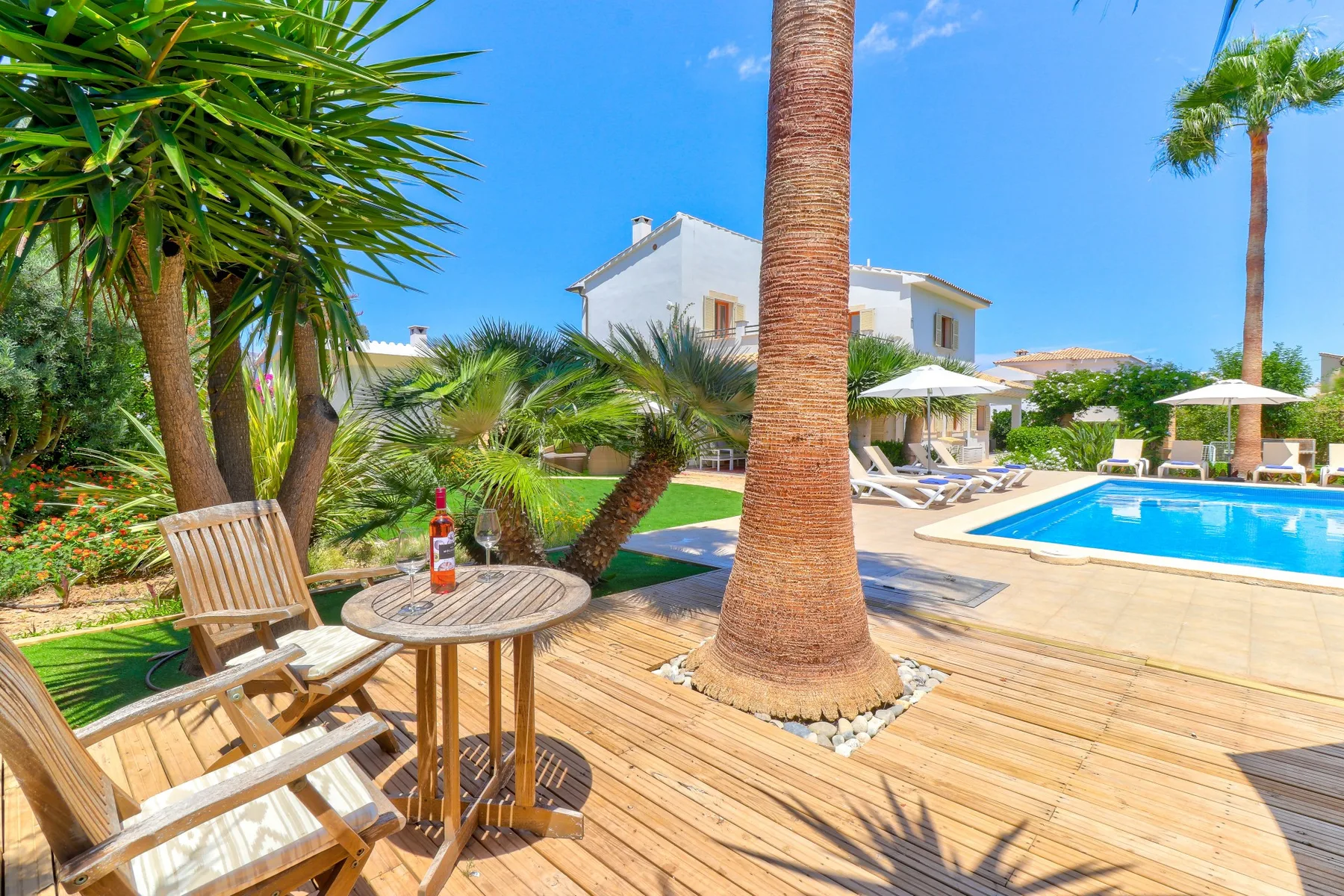 A sunny poolside deck with wooden seating, palm trees, and a white villa.