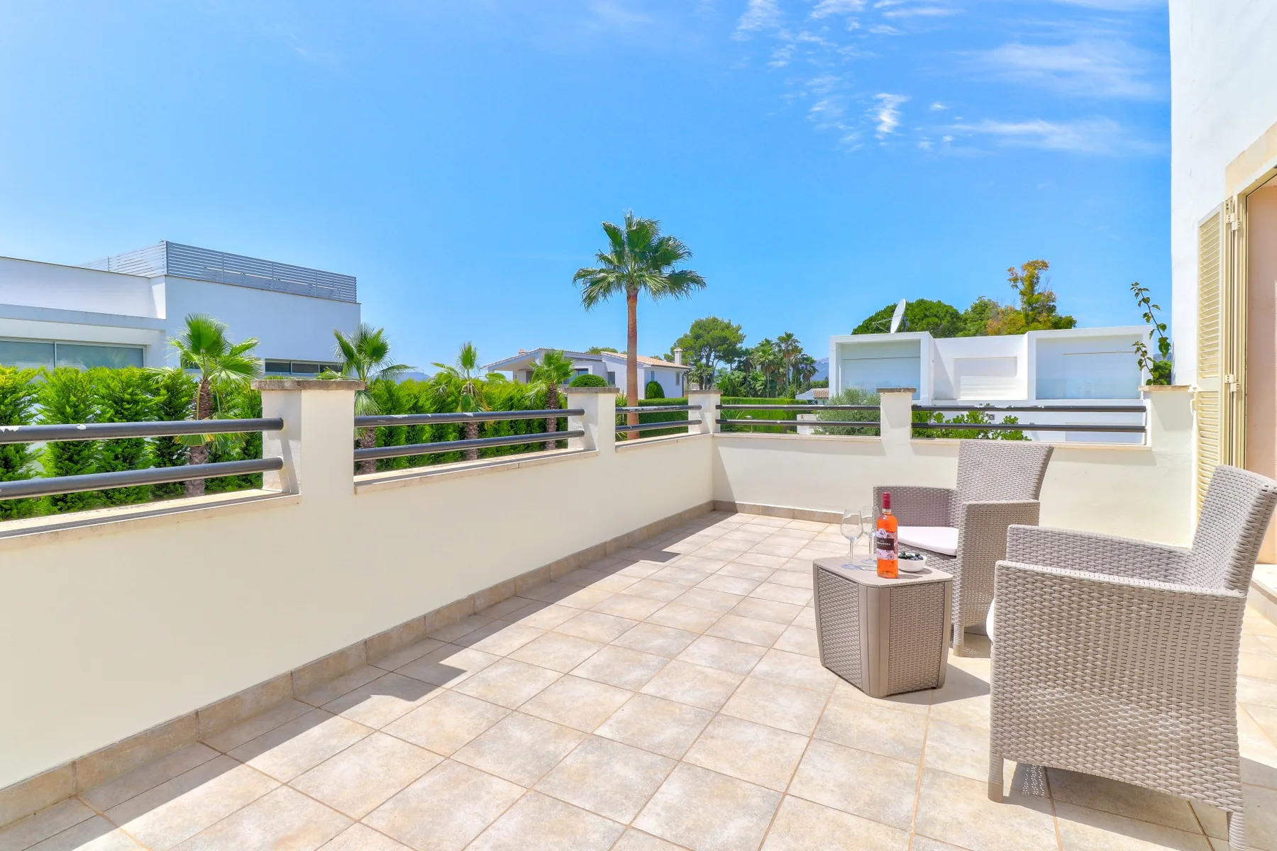 A tiled outdoor terrace has seating and a railing overlooking nearby buildings and greenery.