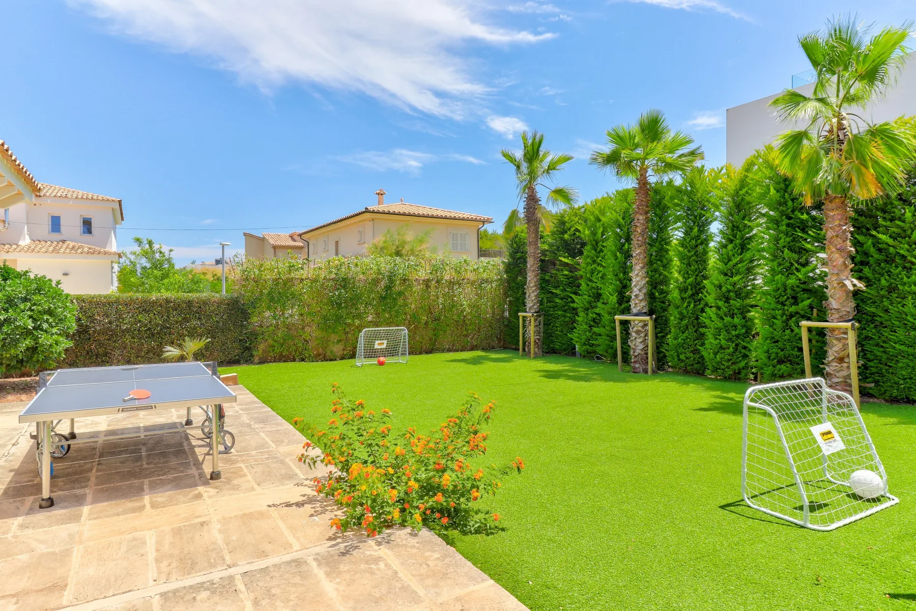A sunny garden with green lawn, palm trees, and a table tennis table.