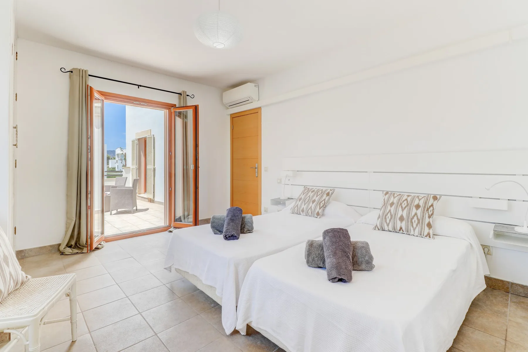 A bright bedroom with two beds and an open glass door leading to a balcony.