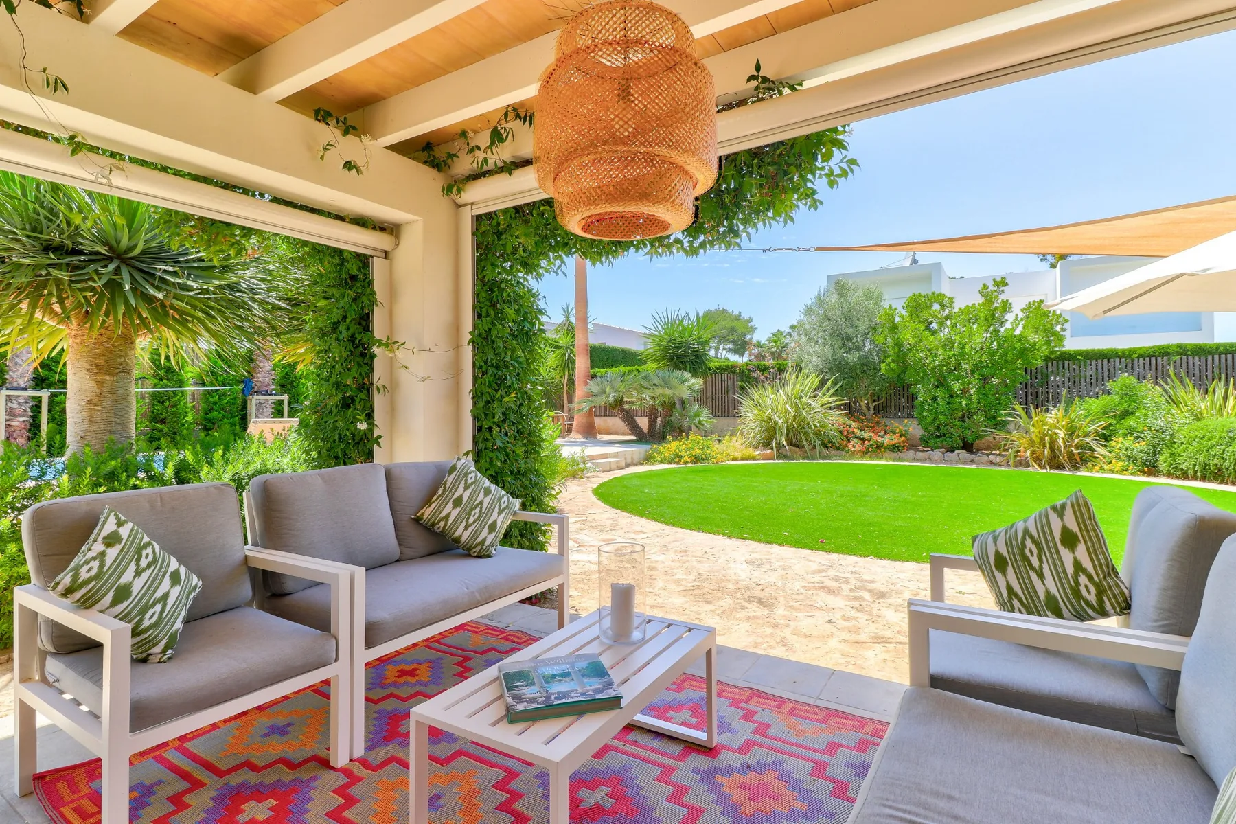 A covered patio with cushioned seating overlooks a lawn and landscaped garden.