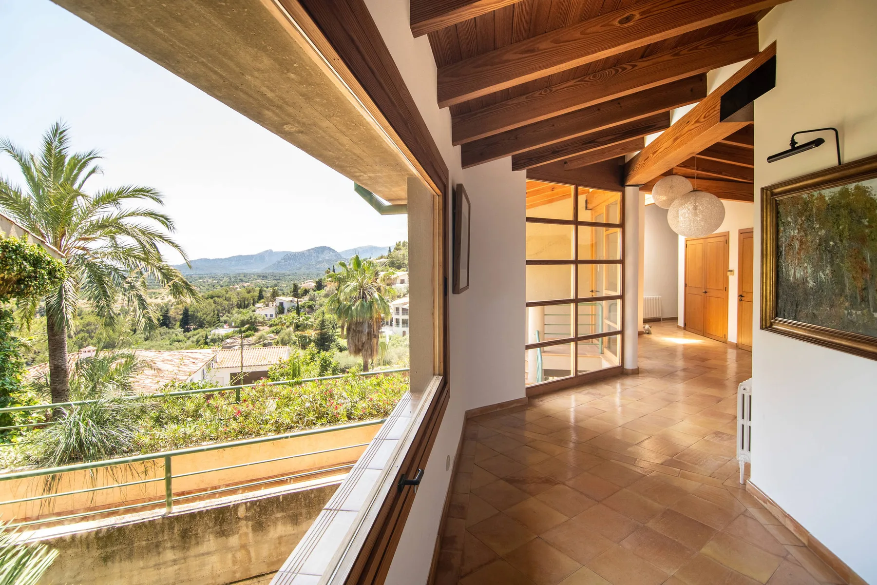 Un pasillo interior luminoso con vigas de madera expuestas y una gran ventana con vistas a la vegetación.