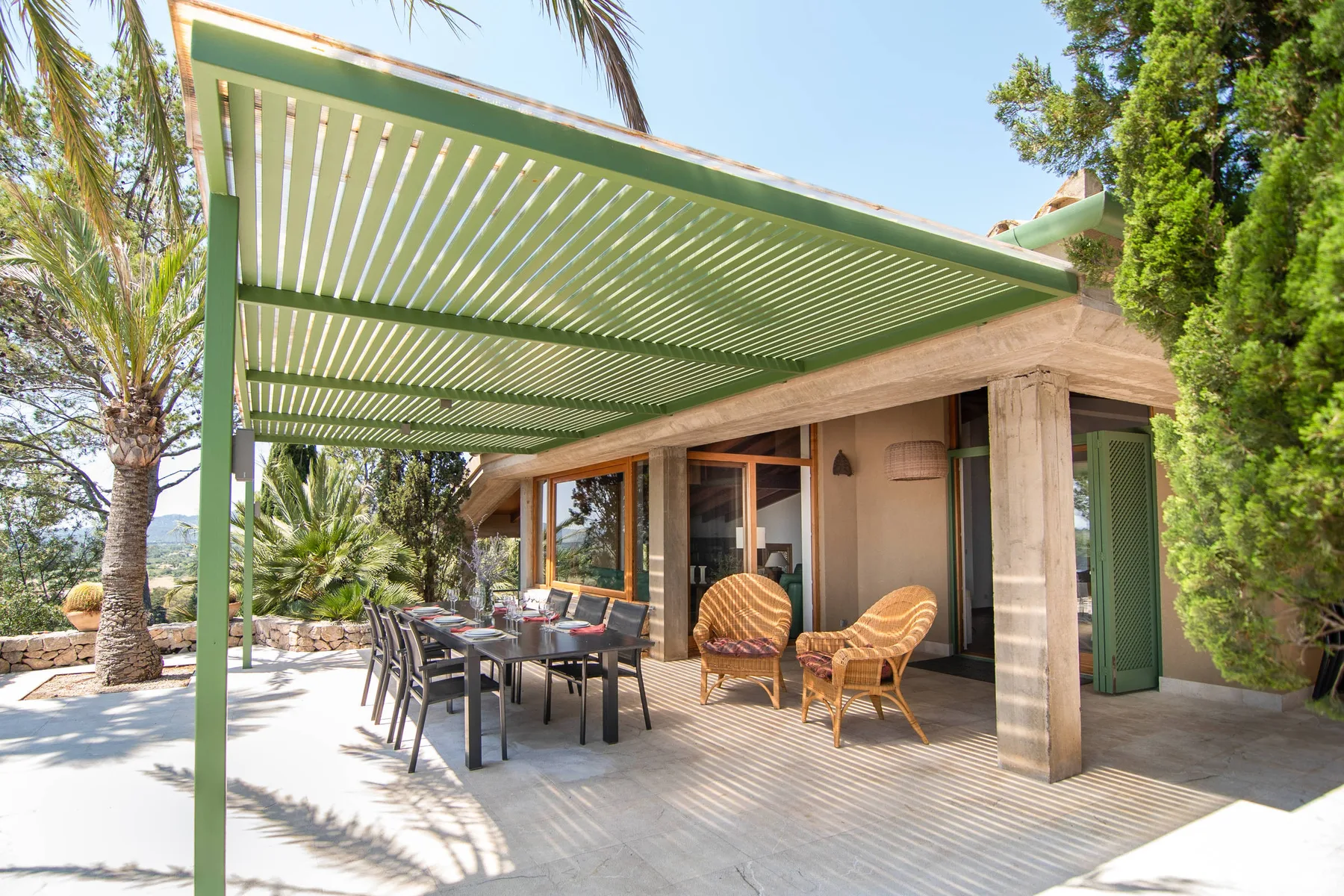 Un patio exterior sombreado con asientos de comedor y sillones de mimbre junto a puertas de vidrio.