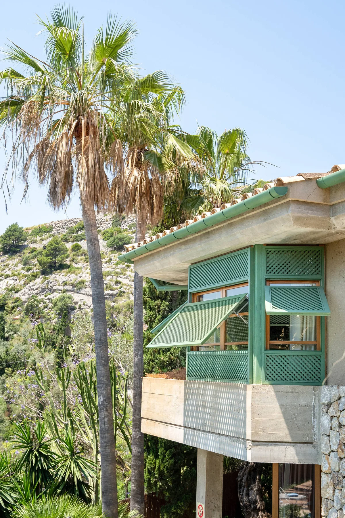 Un edificio elevado con ventanas verdes de celosía está entre palmeras en una ladera soleada.