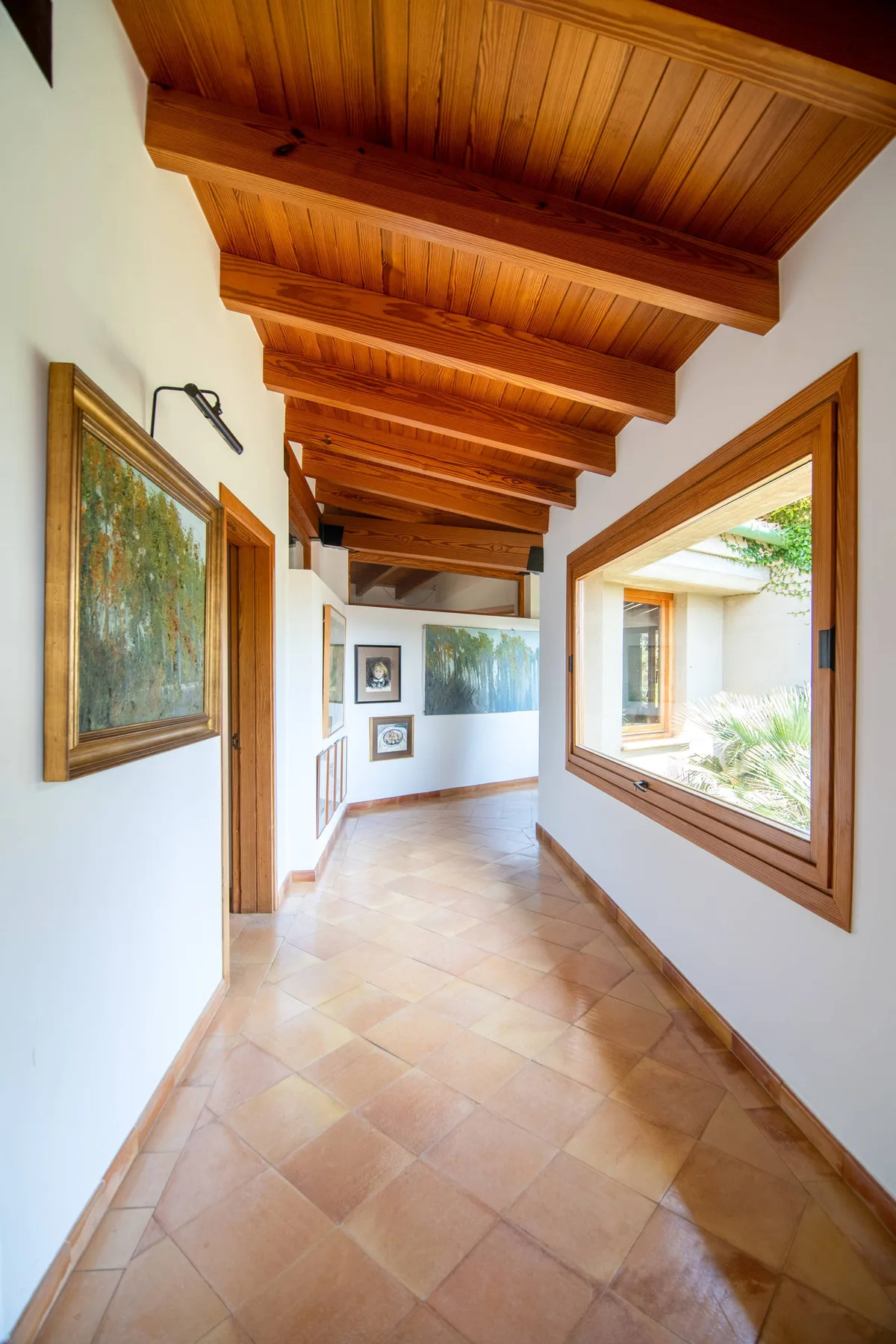 Un pasillo luminoso con suelo de baldosas, vigas de madera vistas y grandes ventanas.