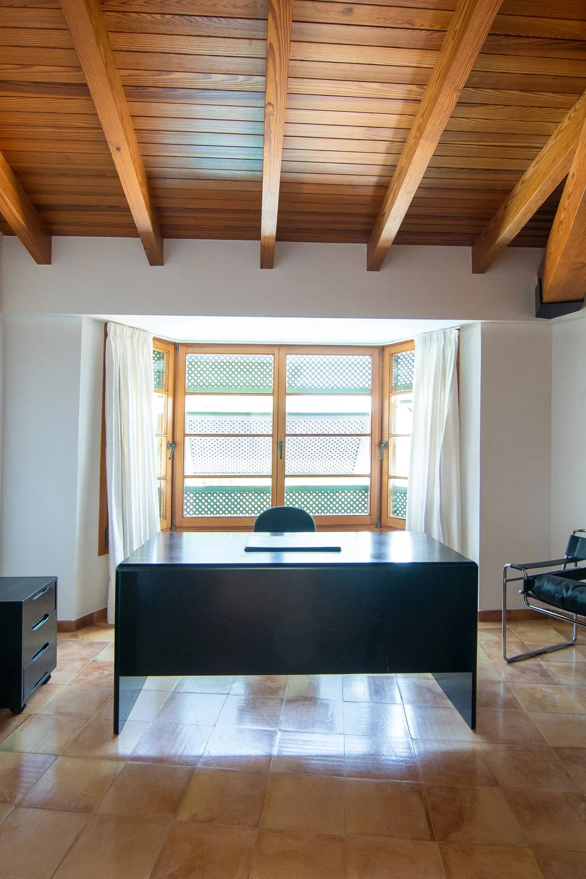 Un escritorio está frente a una ventana con cortinas, bajo vigas de madera expuestas.