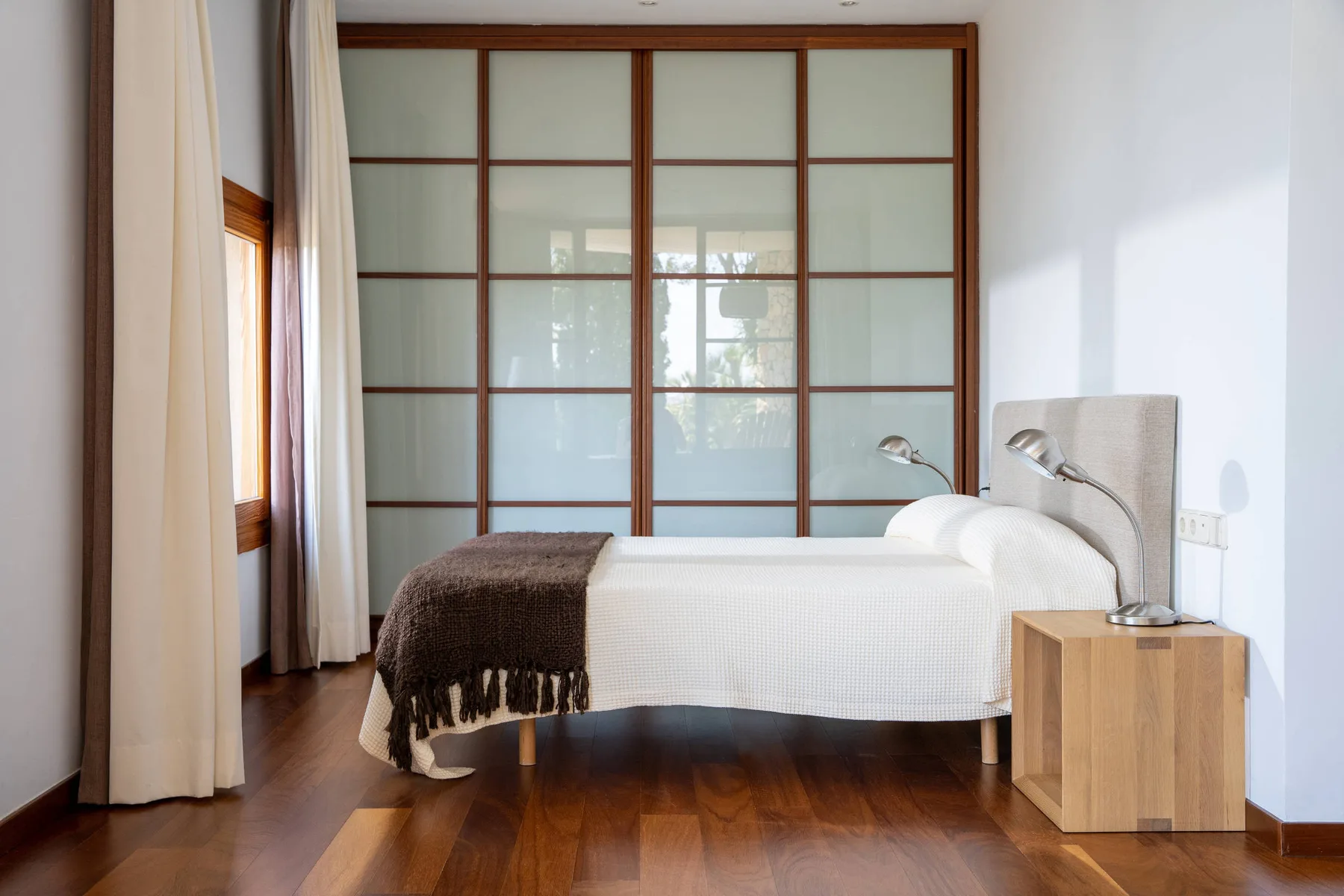 Un dormitorio con una cama bien tendida, una mesita de noche y una ventana amplia con cuadrícula.
